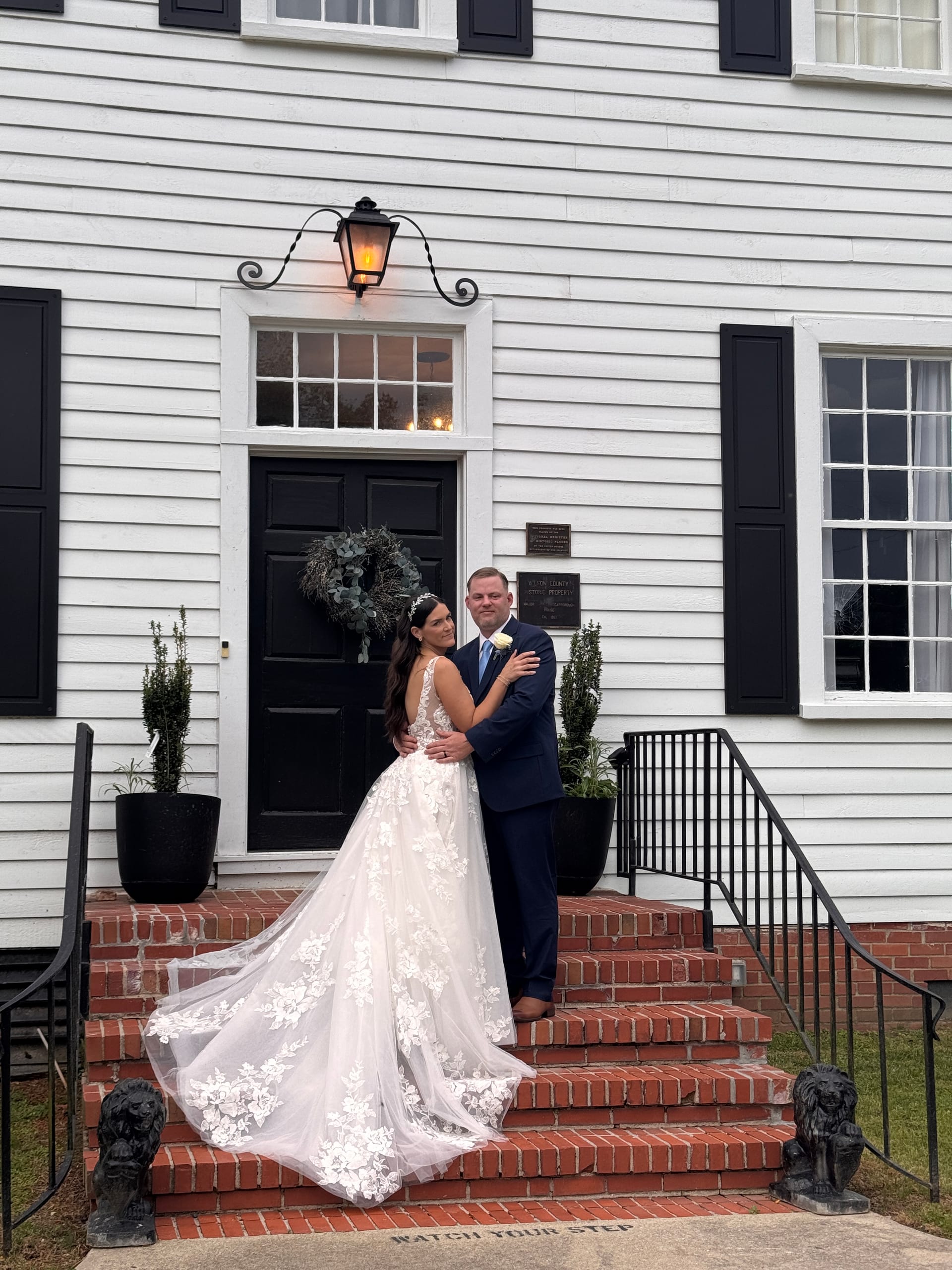 Bride in a floral lace gown with a flowing train and groom in a navy suit embrace on the red brick front steps of the 1821 Scarborough House manor at dusk.
