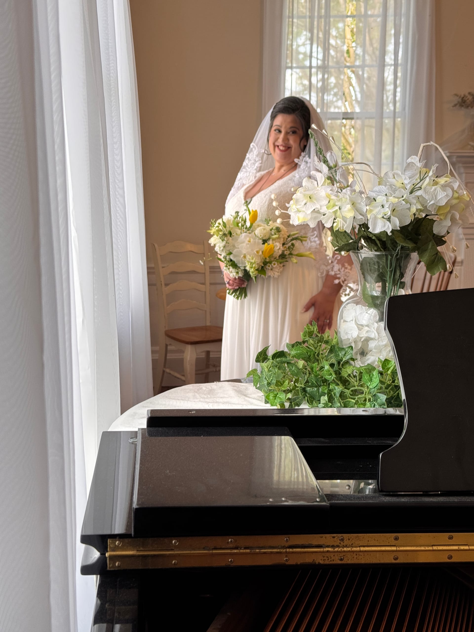 Bride in a white gown and lace veil holding a white and yellow bouquet stands smiling near a window inside the historic grand hall at Scarborough House, with a black grand piano and white floral arrangement in the foreground.