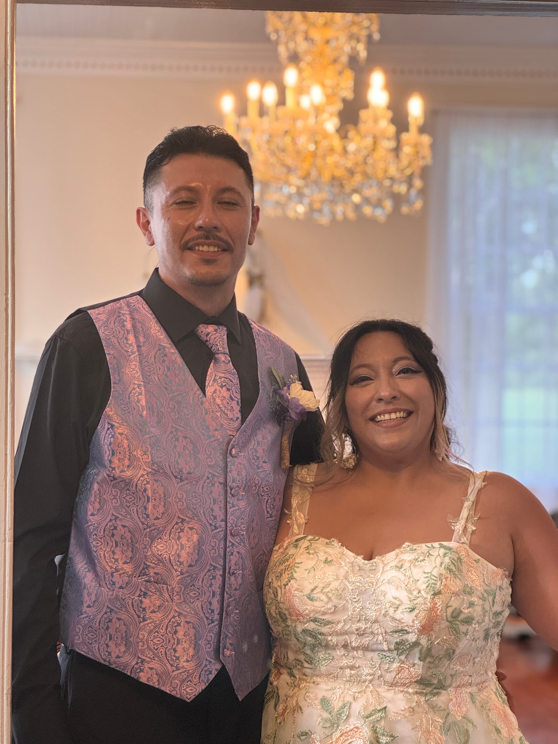 Bride in a floral embroidered gown and groom in a paisley vest and matching tie smile together beneath a crystal chandelier inside the grand hall of Scarborough House.