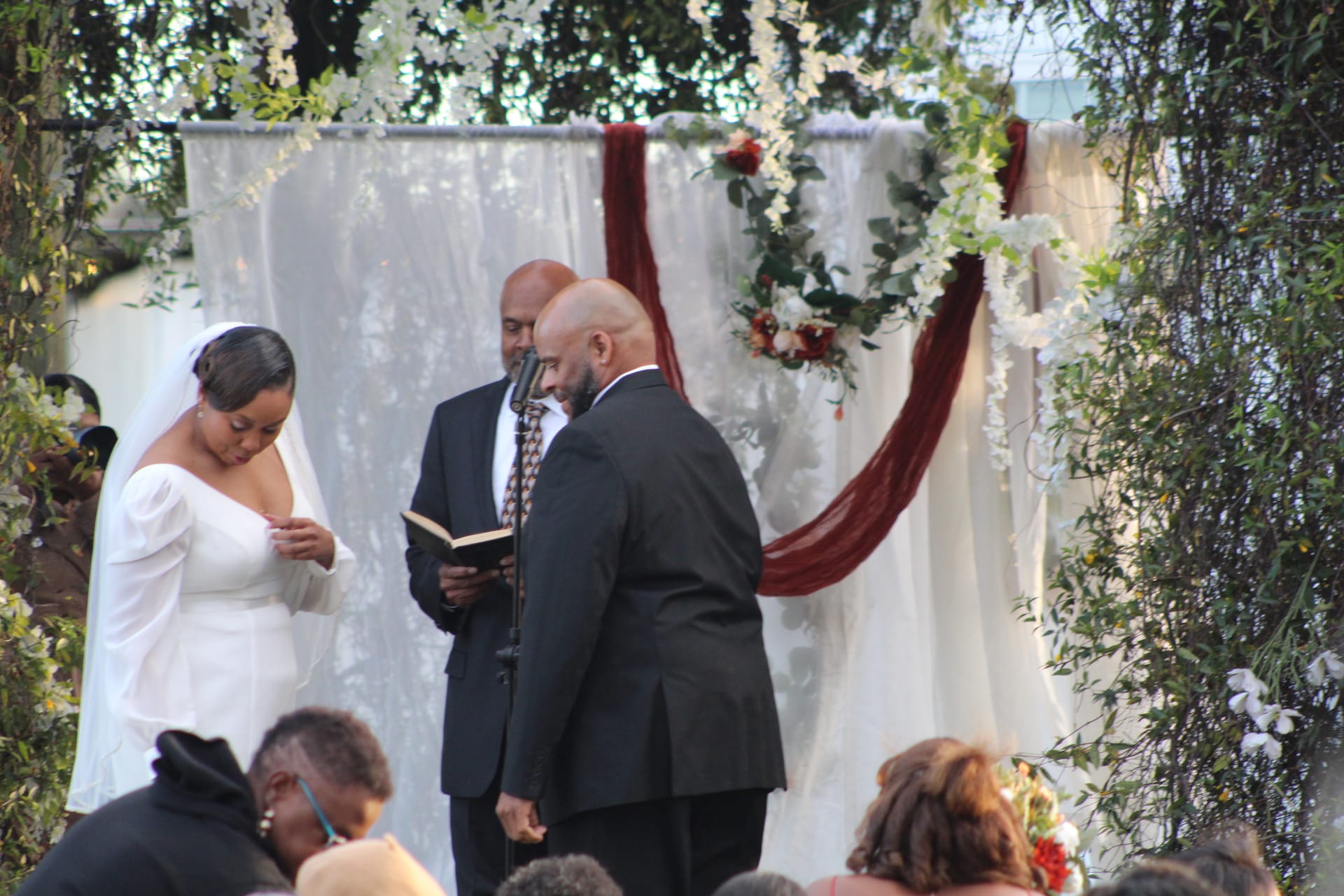 Bride in a white off-shoulder gown reads her vows to the groom before an officiant beneath a draped floral arch adorned with white flowers and burgundy fabric at Scarborough House.