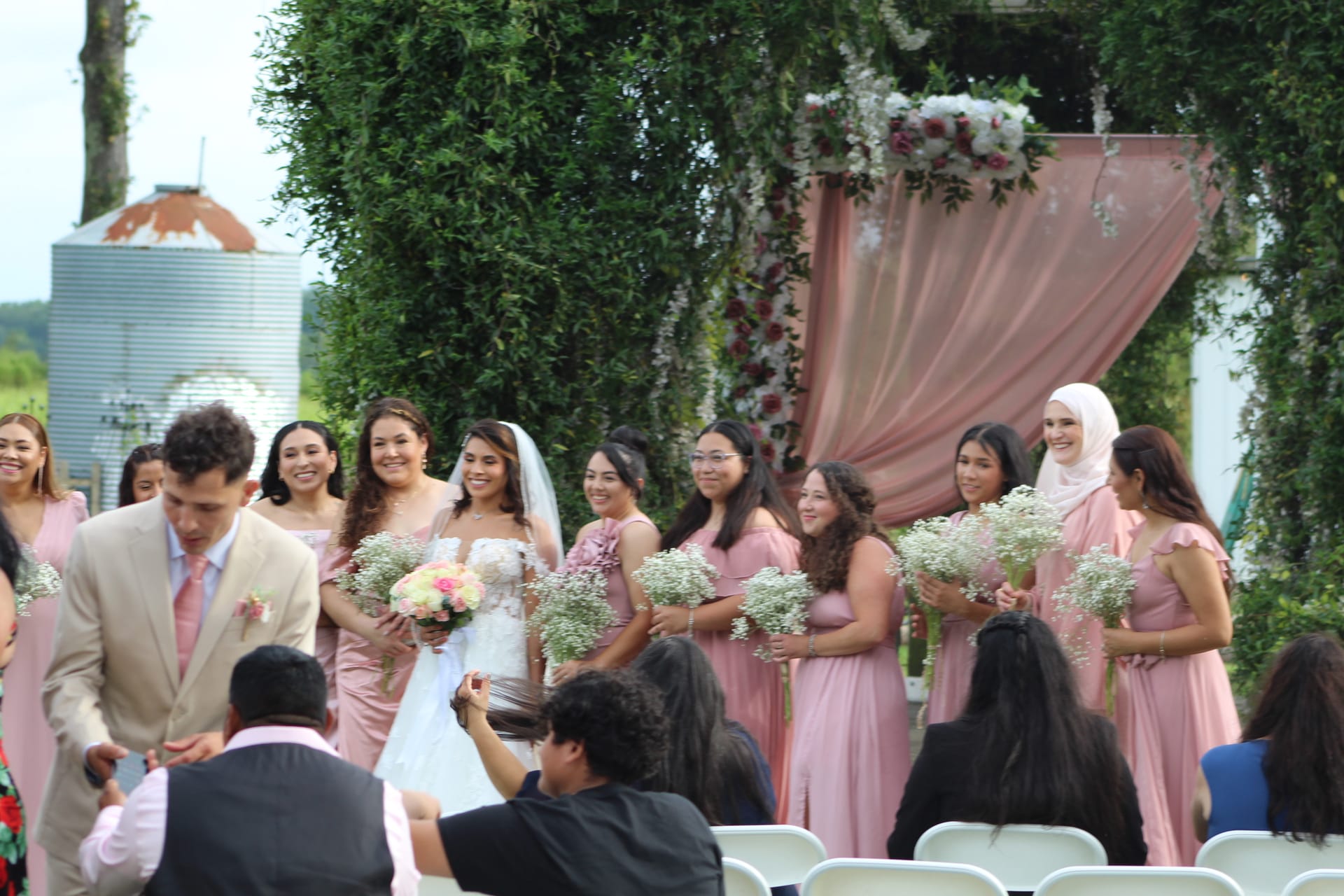 Smiling bride in white gown and veil walks with her bridesmaids in blush pink dresses holding bouquets past a floral-draped arch at an outdoor wedding ceremony.