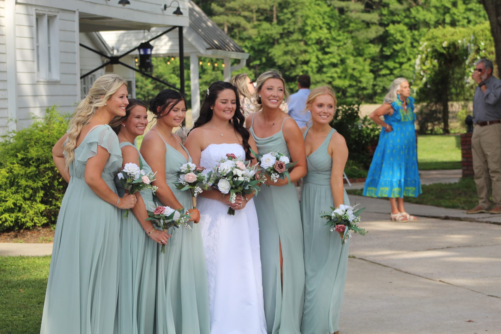 Smiling bride in a white strapless gown holds a white and blush bouquet surrounded by six bridesmaids in sage green dresses outside a historic white manor house.