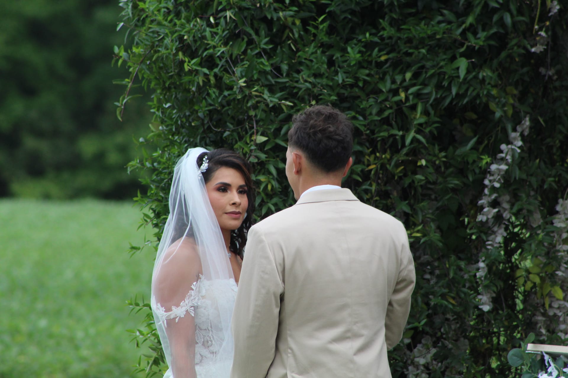 Bride in a lace gown and flowing veil gazes at her groom during an outdoor wedding ceremony at Scarborough House, framed by a lush green hedge backdrop.