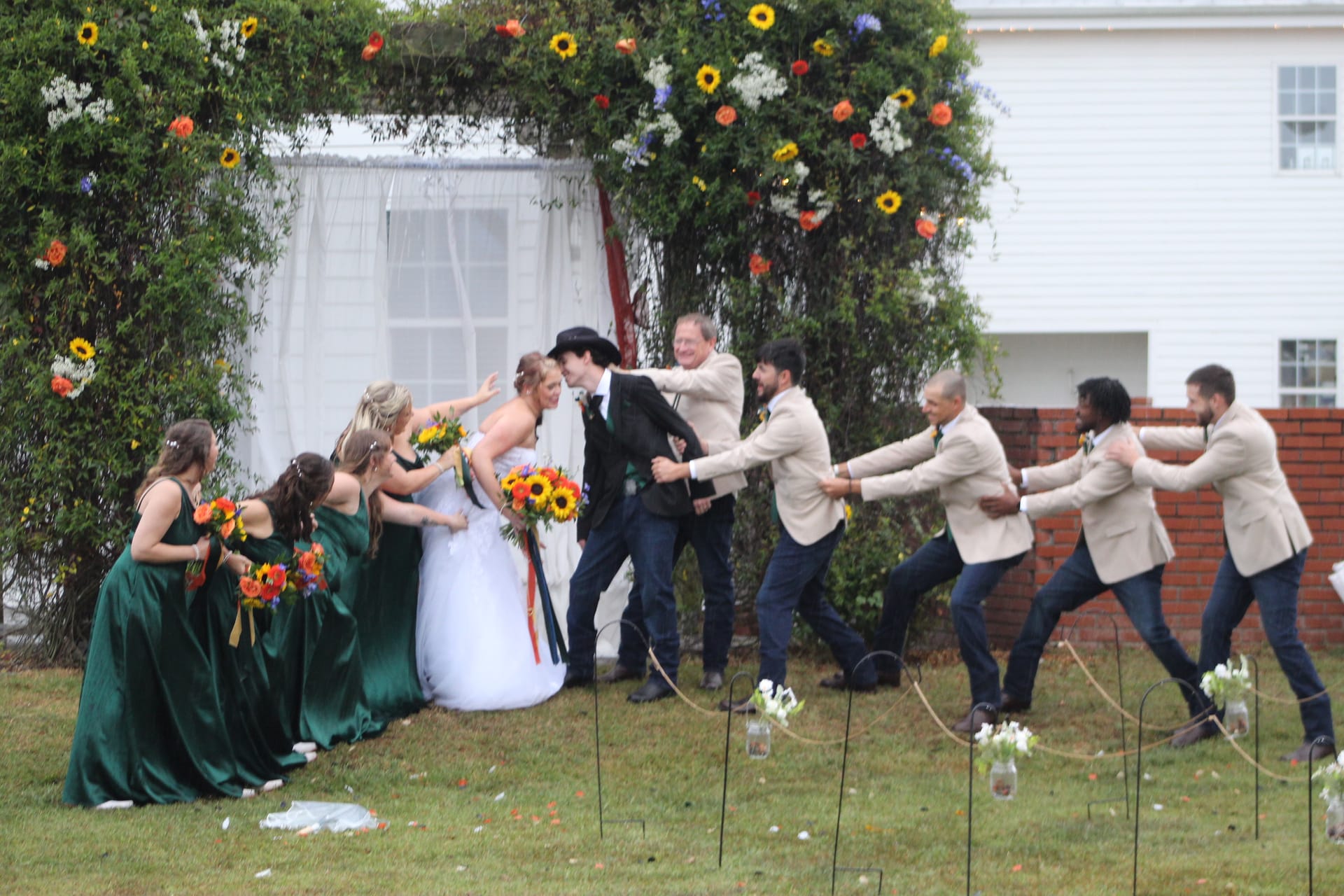 Bride and groom share a kiss while bridesmaids in emerald green and groomsmen in tan blazers playfully pull them apart on the ceremony lawn beneath a sunflower-adorned floral arch at Scarborough House.