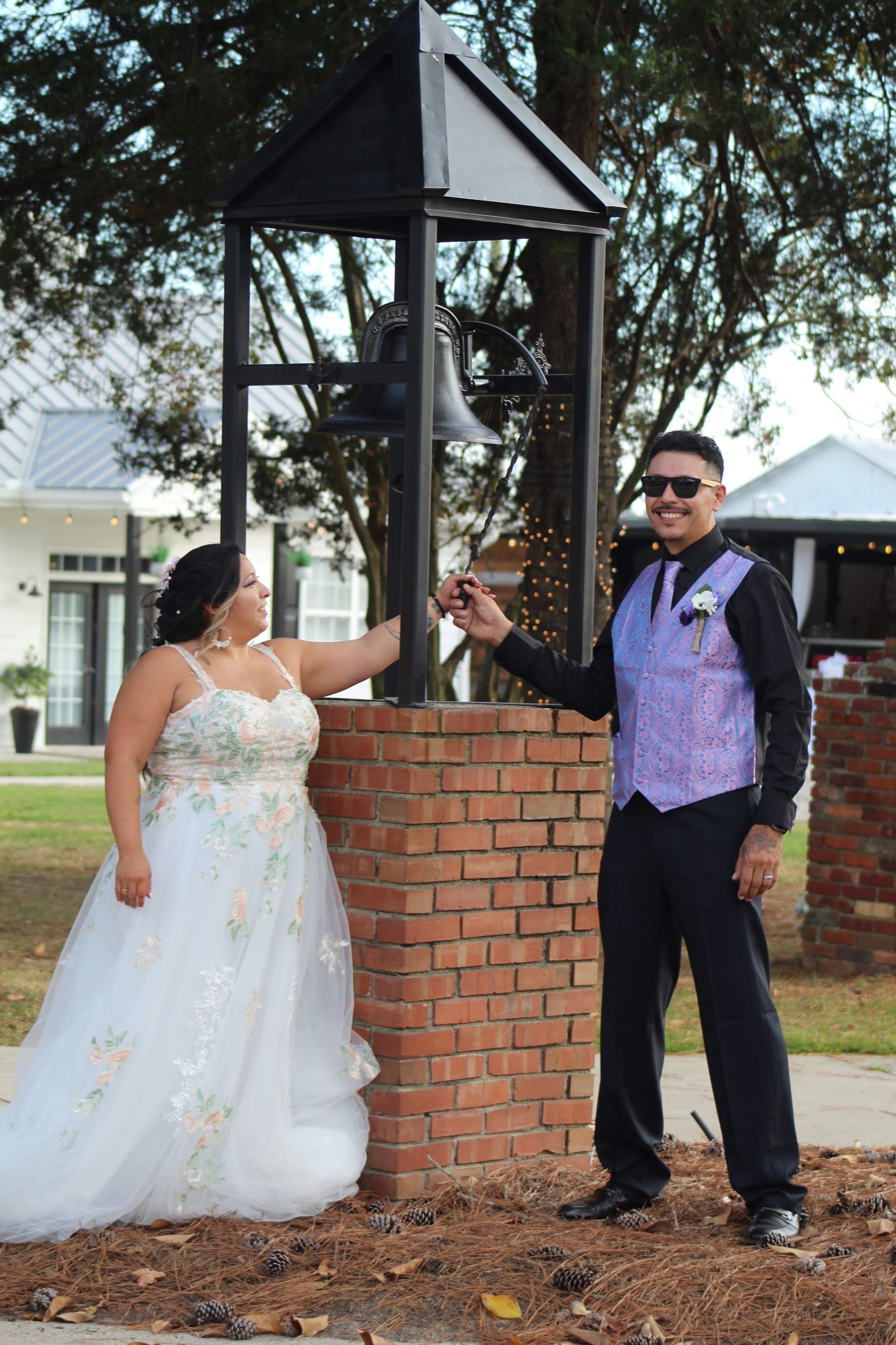 Bride in a floral appliqué gown and groom in a purple vest and black shirt ring the decorative bell together on the ceremony grounds of Scarborough House wedding venue.