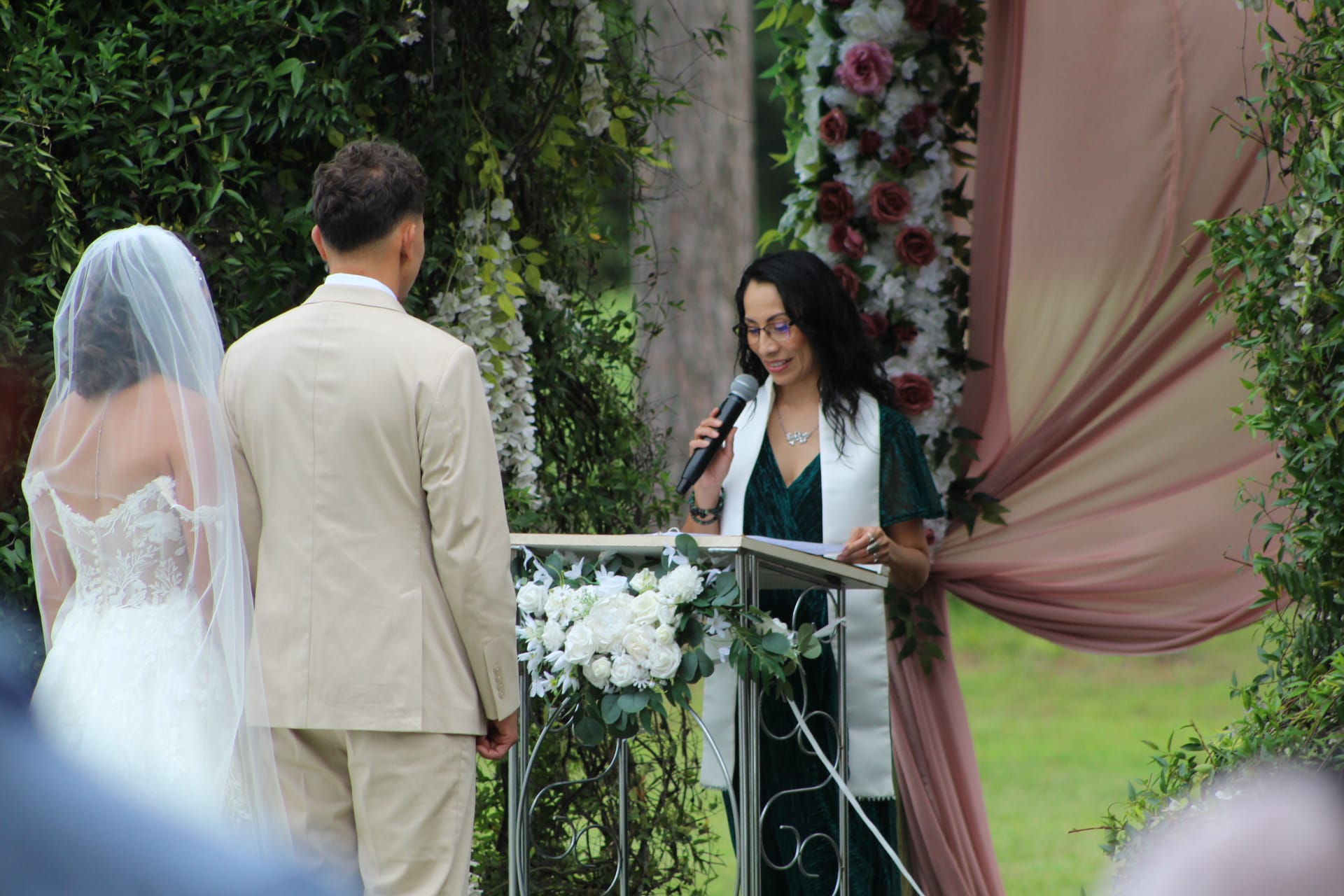 Wedding officiant speaks into a microphone at a floral-adorned podium as a bride in a lace gown and groom in a tan suit stand before her during an outdoor garden ceremony at Scarborough House.