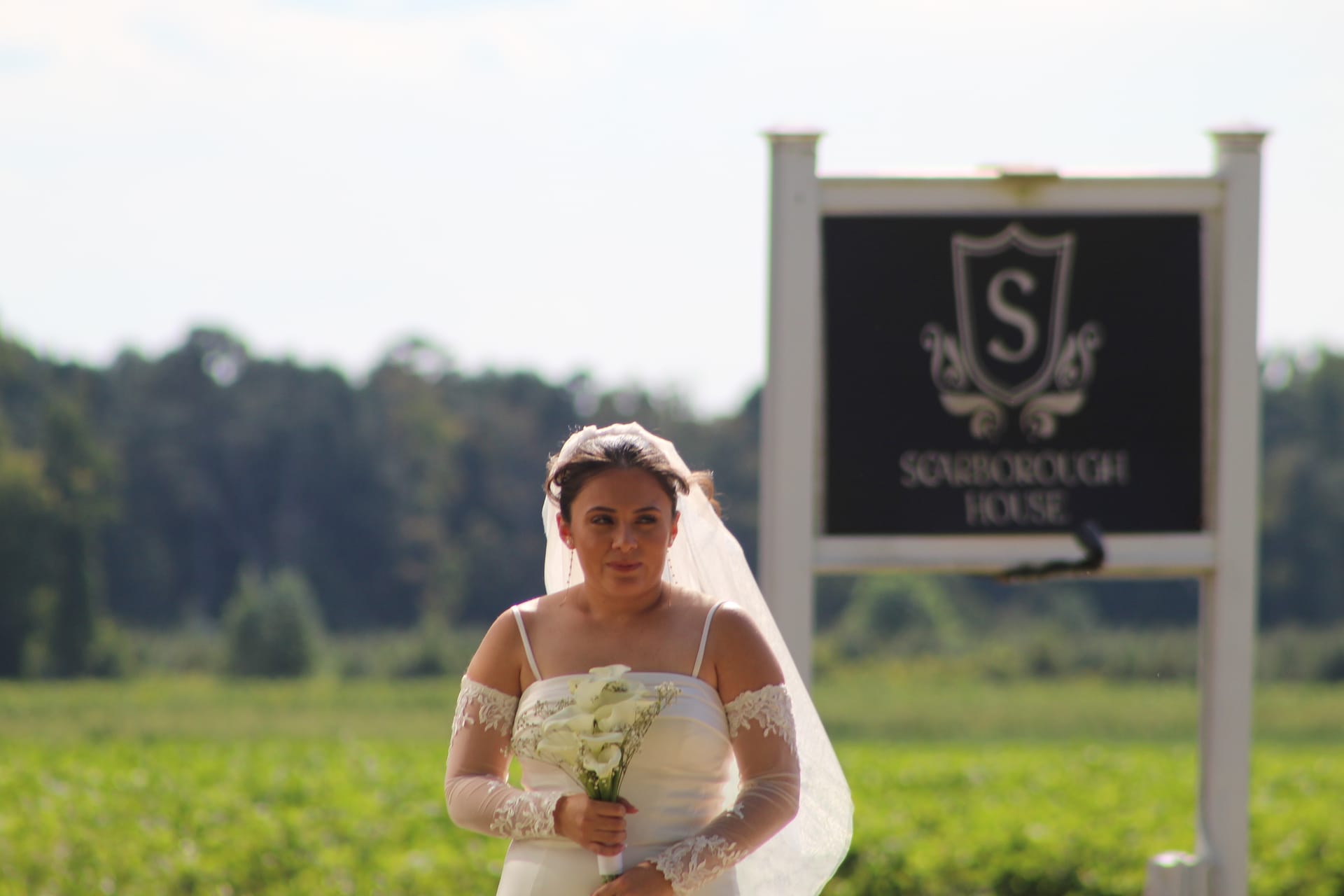 Bride in a lace off-shoulder wedding gown holding a white bouquet stands before the Scarborough House marquee sign surrounded by green fields.