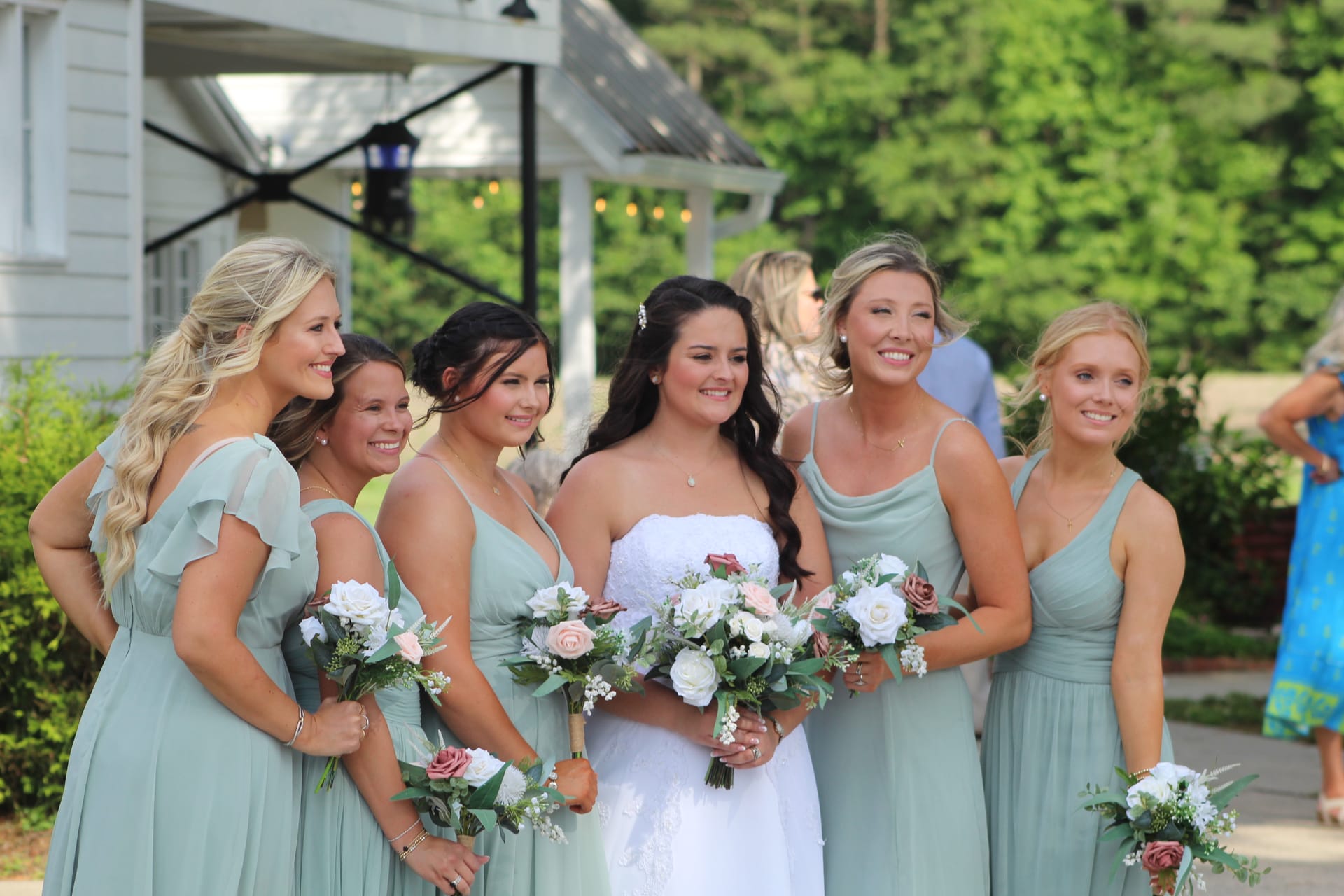 Bride in a white strapless gown stands surrounded by five bridesmaids in sage green dresses holding white and blush floral bouquets outside the historic Scarborough House manor.