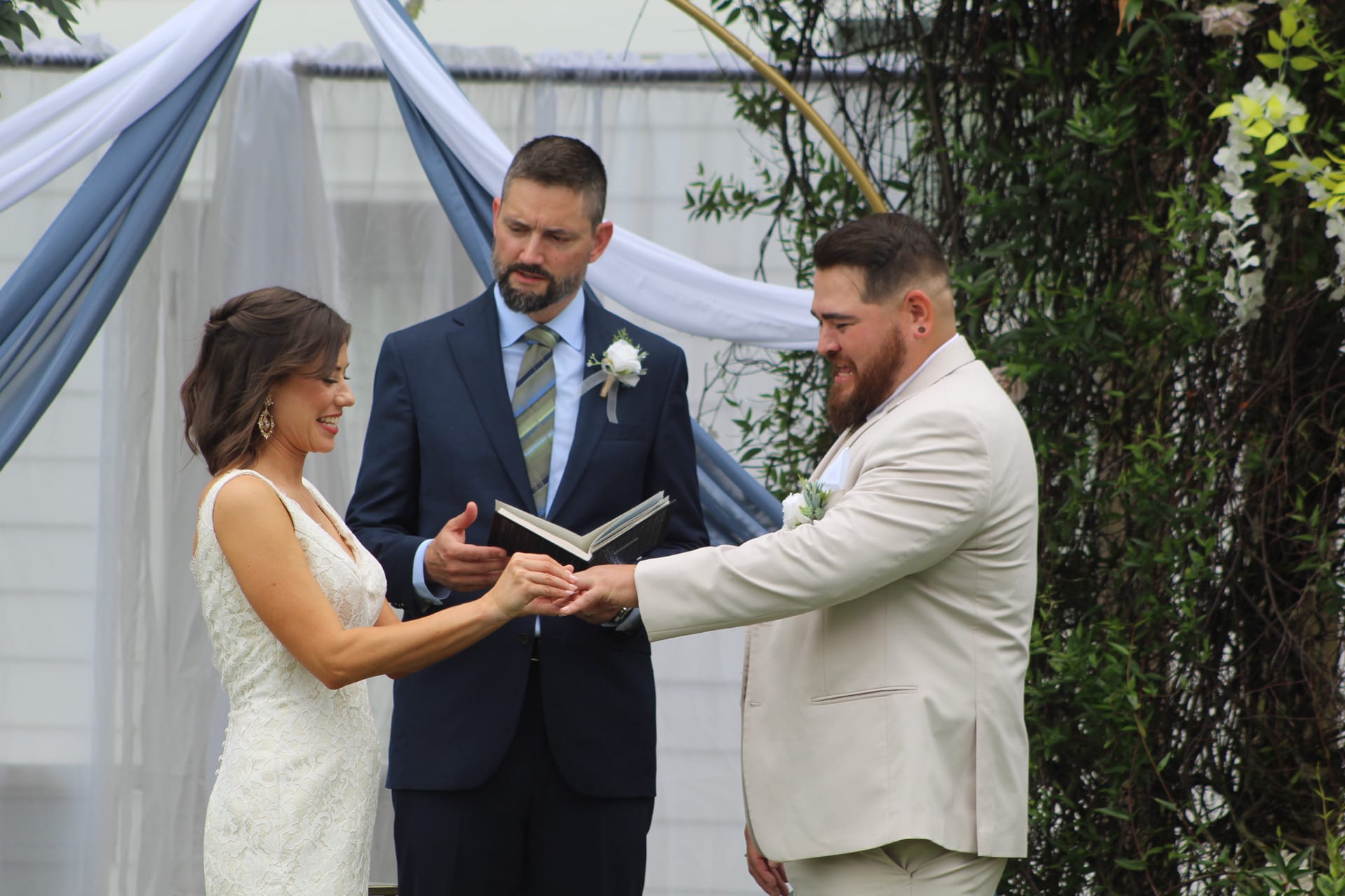 Bride and groom exchange rings during an outdoor wedding ceremony beneath a floral and greenery arch with blue draped fabric at Scarborough House.