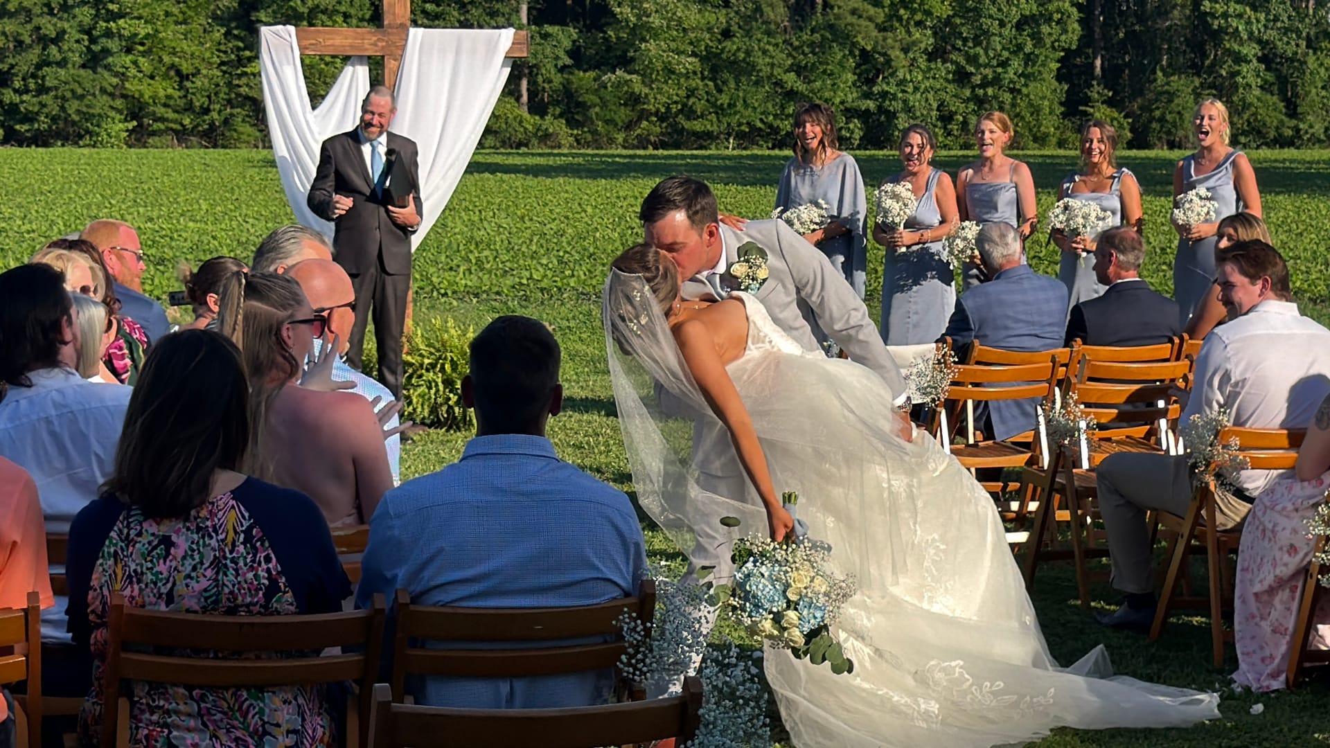 Groom dips his bride for their first kiss during an outdoor ceremony on the sunlit lawn at Scarborough House, surrounded by bridesmaids in dusty blue and smiling guests seated in wooden chairs.