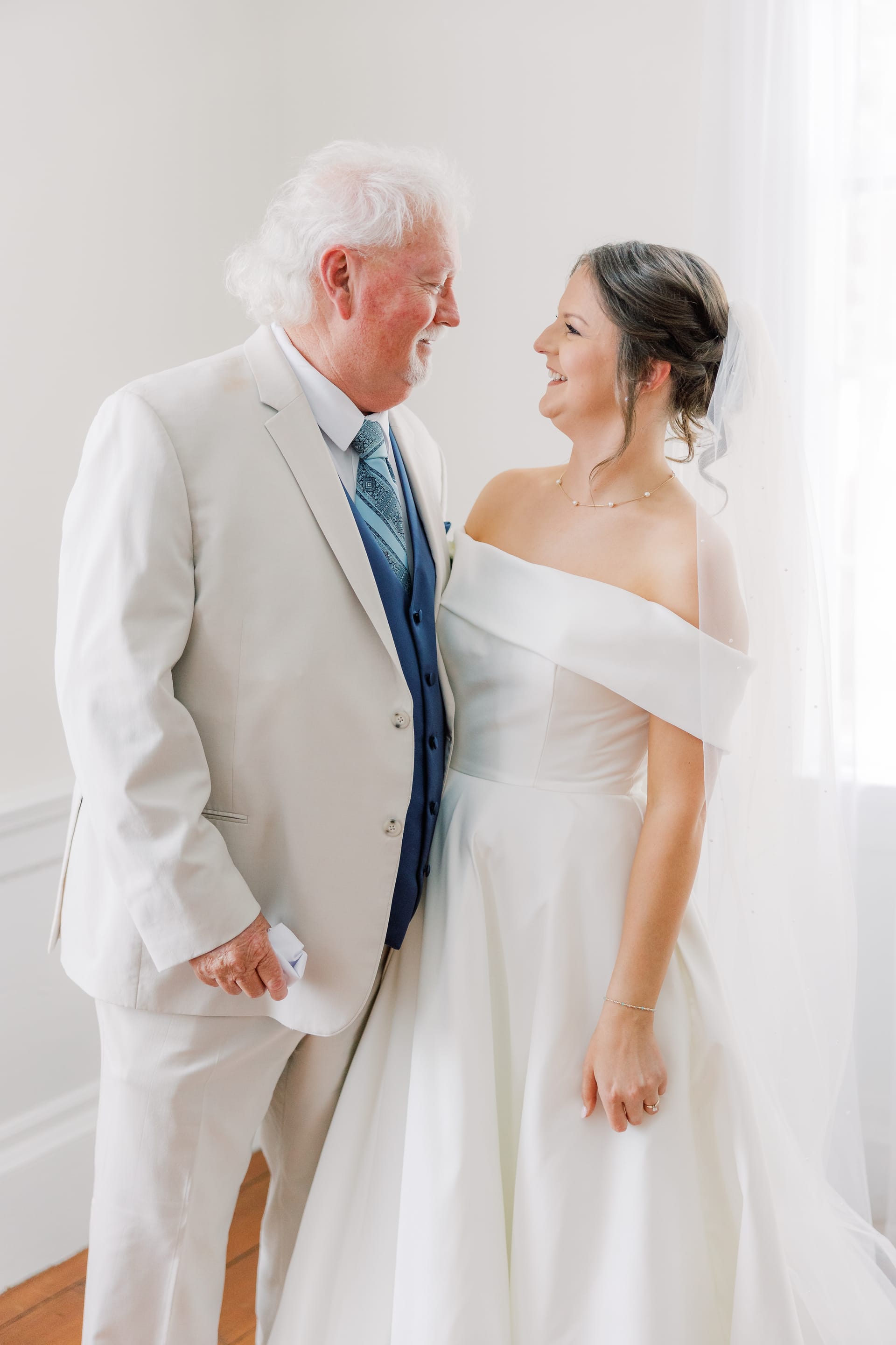 A bride in an off-the-shoulder white gown and veil shares a joyful smile with an older gentleman in a cream suit and blue vest inside Scarborough House.