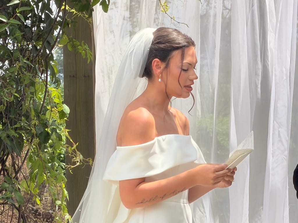 Bride in an off-shoulder white gown and veil reads her handwritten vows during an outdoor garden ceremony at Scarborough House historic estate.
