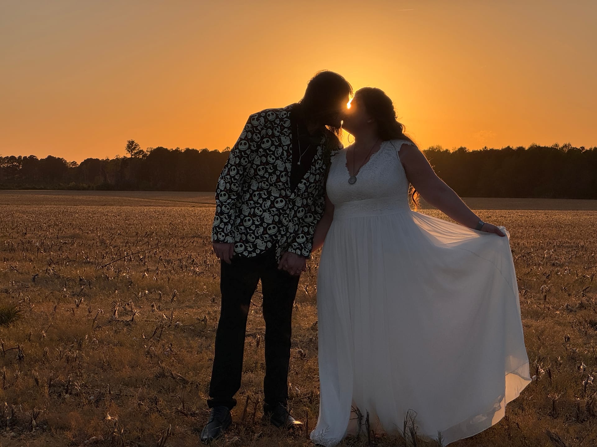 Bride in a flowing white gown and groom in a black-and-white patterned blazer share a kiss silhouetted against a glowing orange sunset over the open farmland fields at Scarborough House.