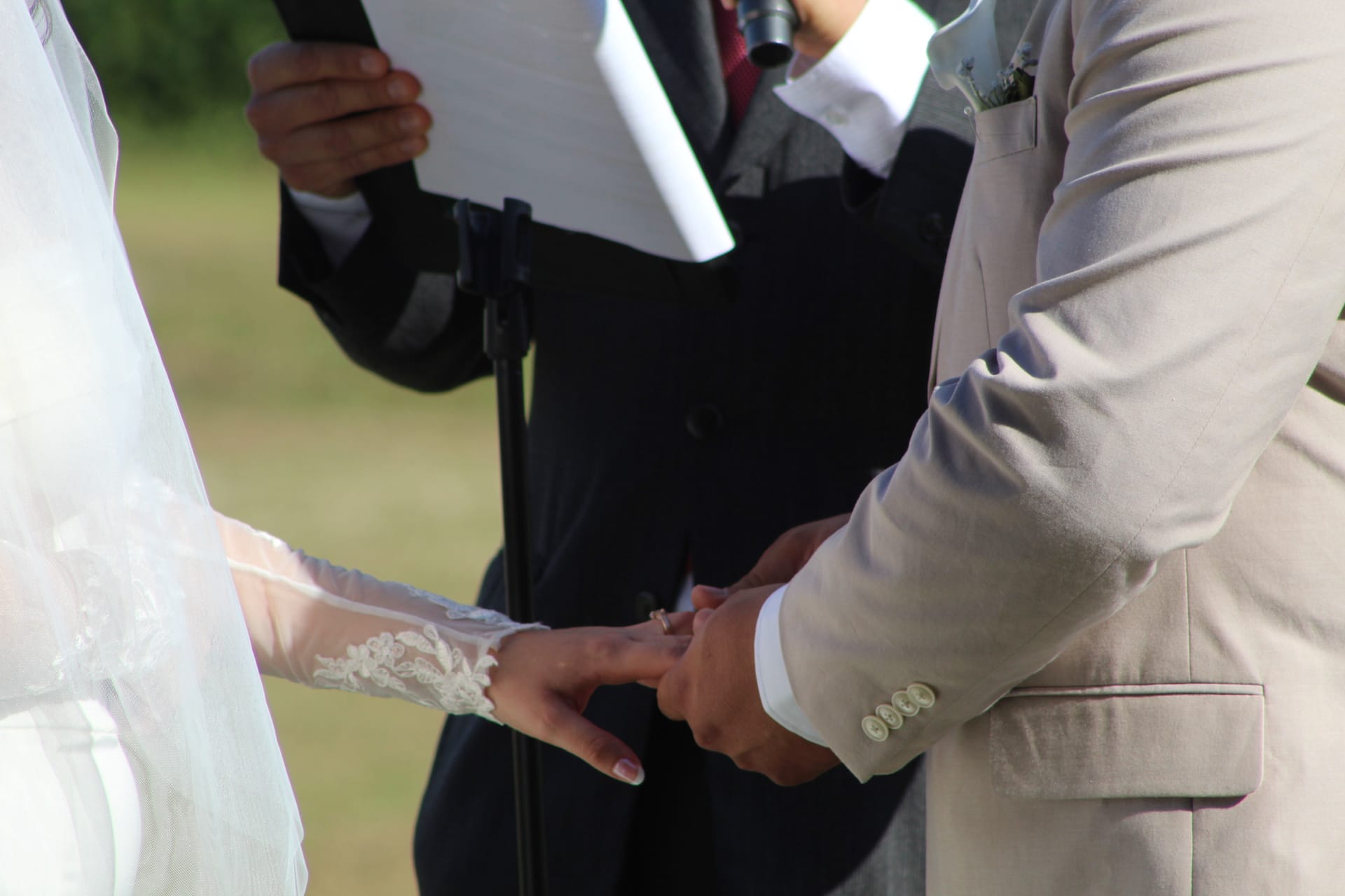Groom in a tan suit places a wedding ring on the bride's lace-gloved hand during an outdoor ceremony at Scarborough House as the officiant reads from papers in the background.