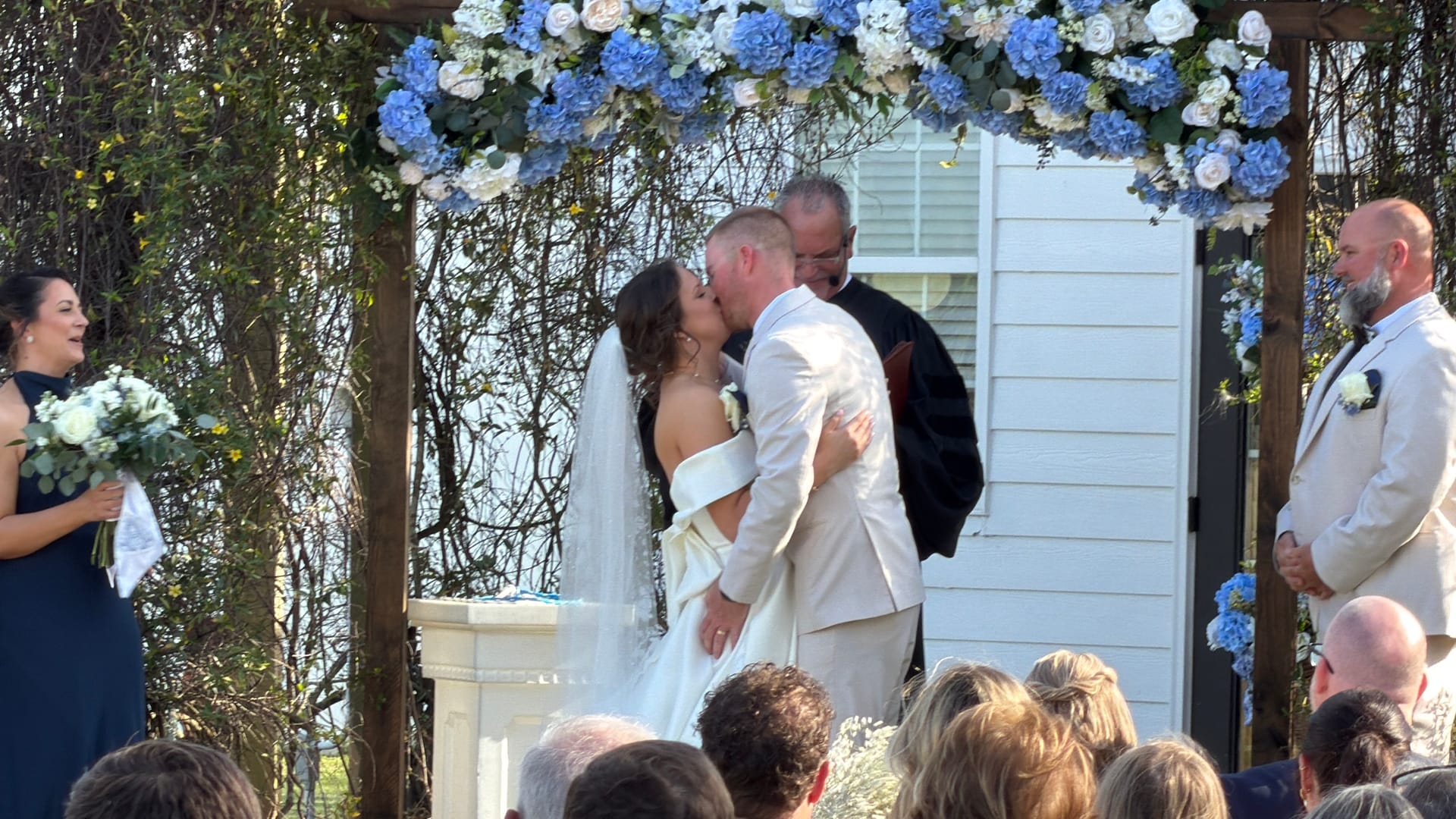 Bride and groom share their first kiss under an elegant blue hydrangea and white rose floral arch during an outdoor wedding ceremony at Scarborough House.
