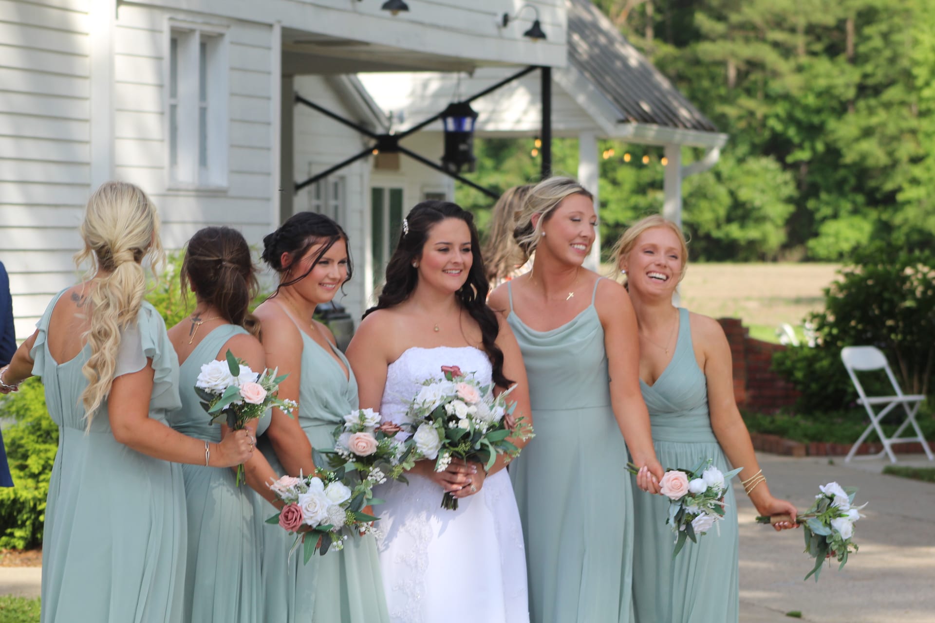 Bride in white gown laughing with five bridesmaids in sage green dresses holding pink and white bouquets outside the historic Scarborough House manor.