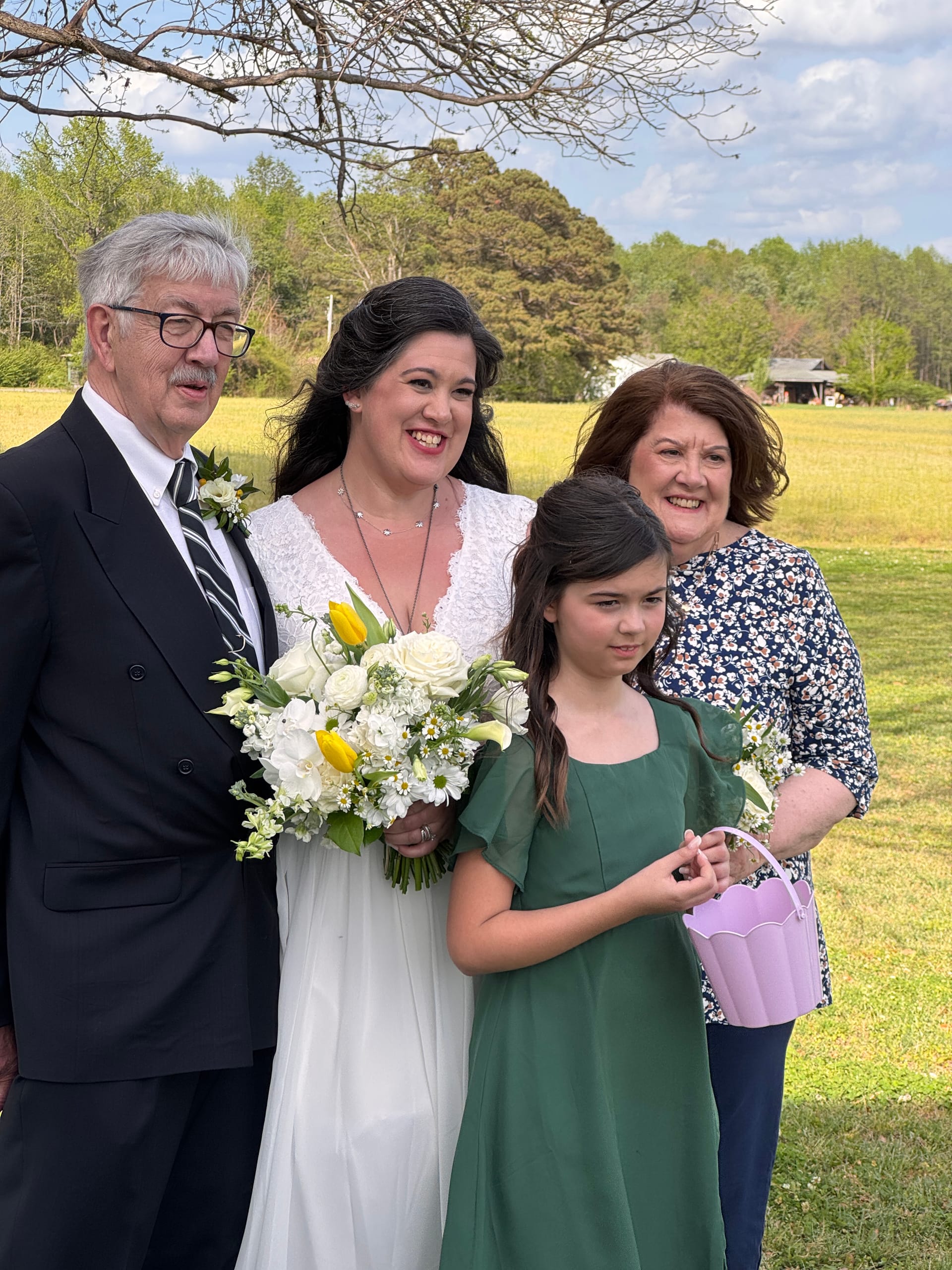 Bride in white lace gown holding a yellow and white bouquet poses with her parents and a young flower girl in a green dress against a sunny farmland field backdrop.