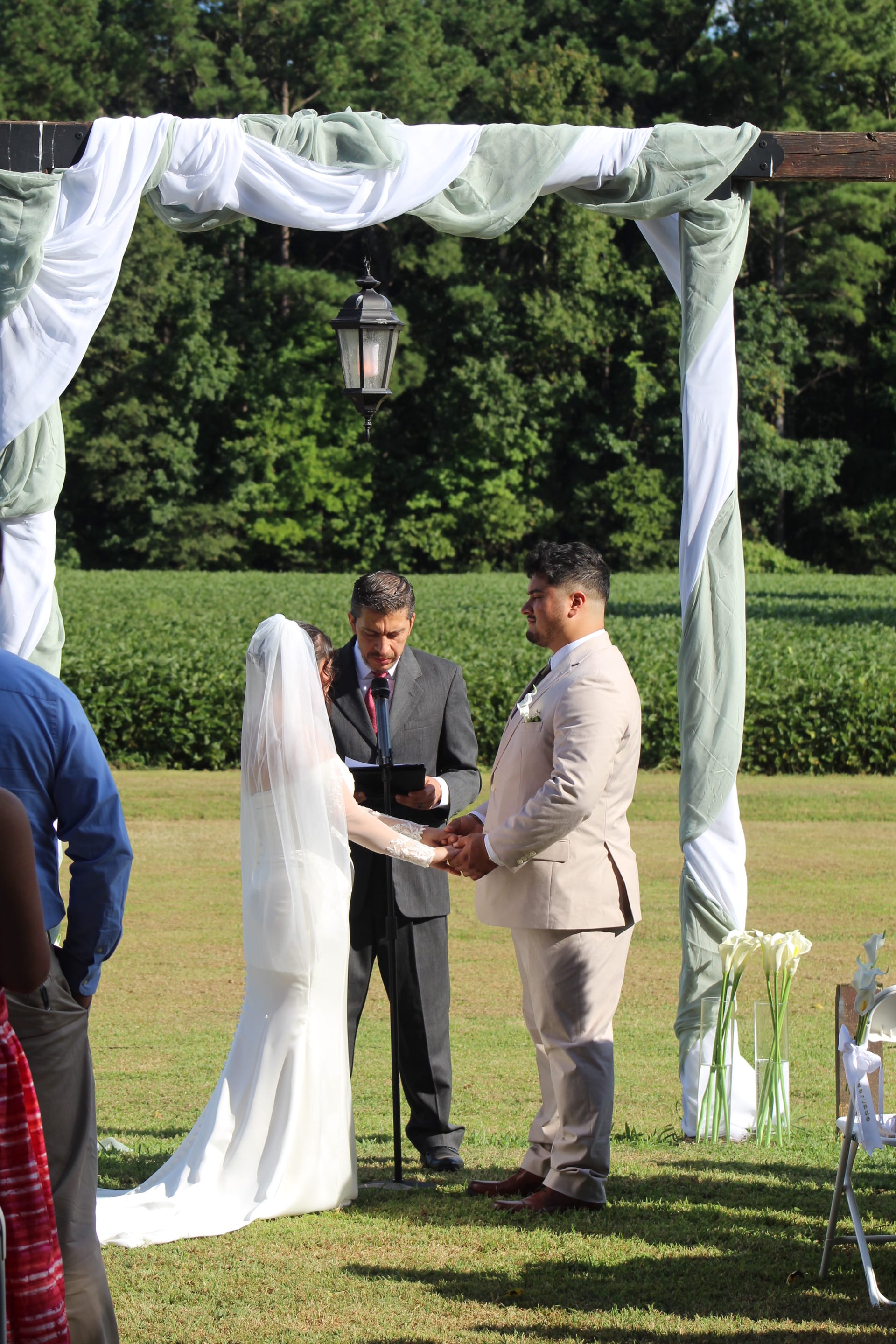Bride and groom exchange vows beneath a draped wooden arbor on the ceremony lawn at Scarborough House, surrounded by lush green fields and trees.