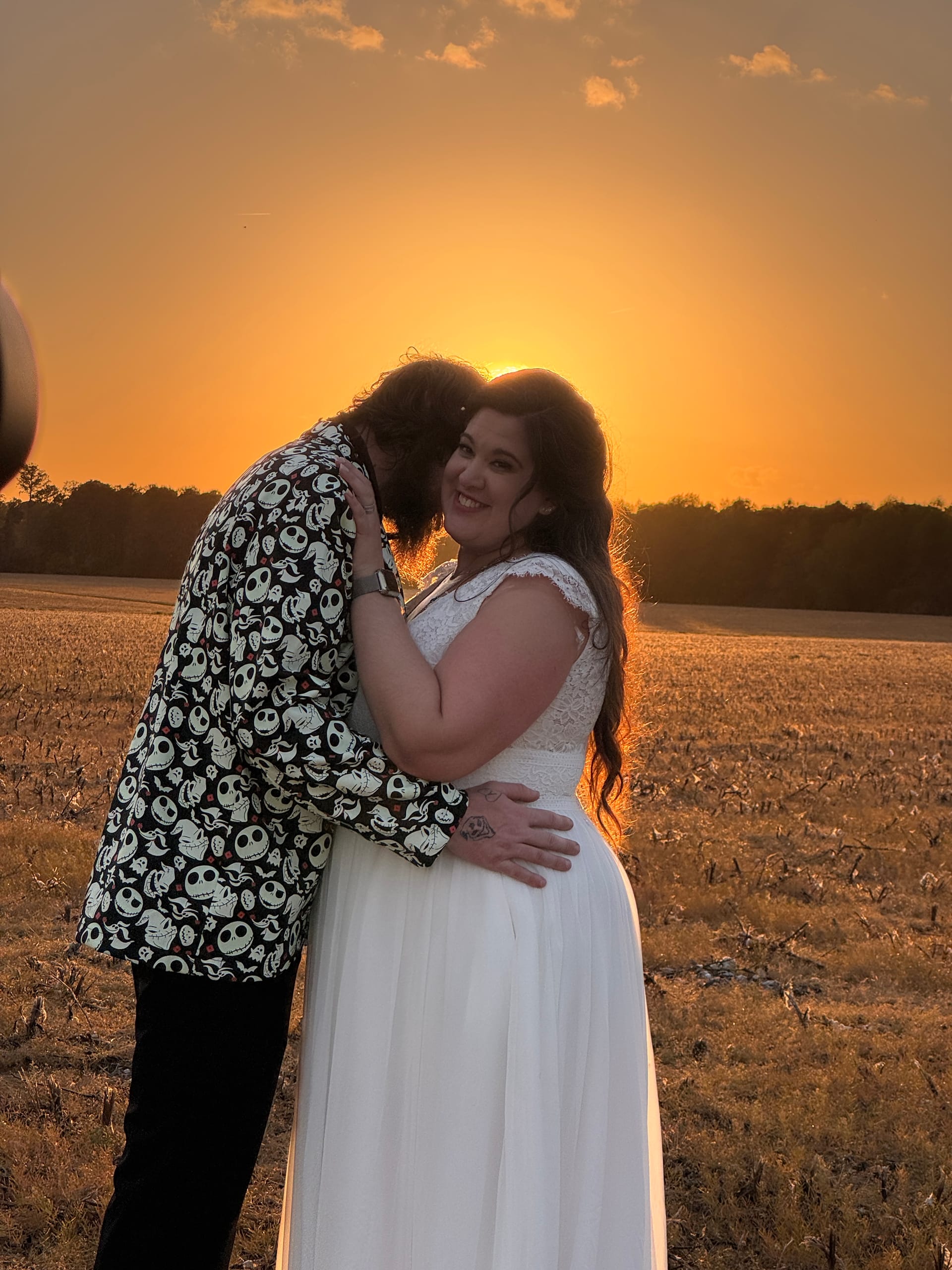 Bride in white lace gown smiles warmly as groom in a Nightmare Before Christmas printed jacket embraces her during a vivid golden sunset over the open farmland fields at Scarborough House.
