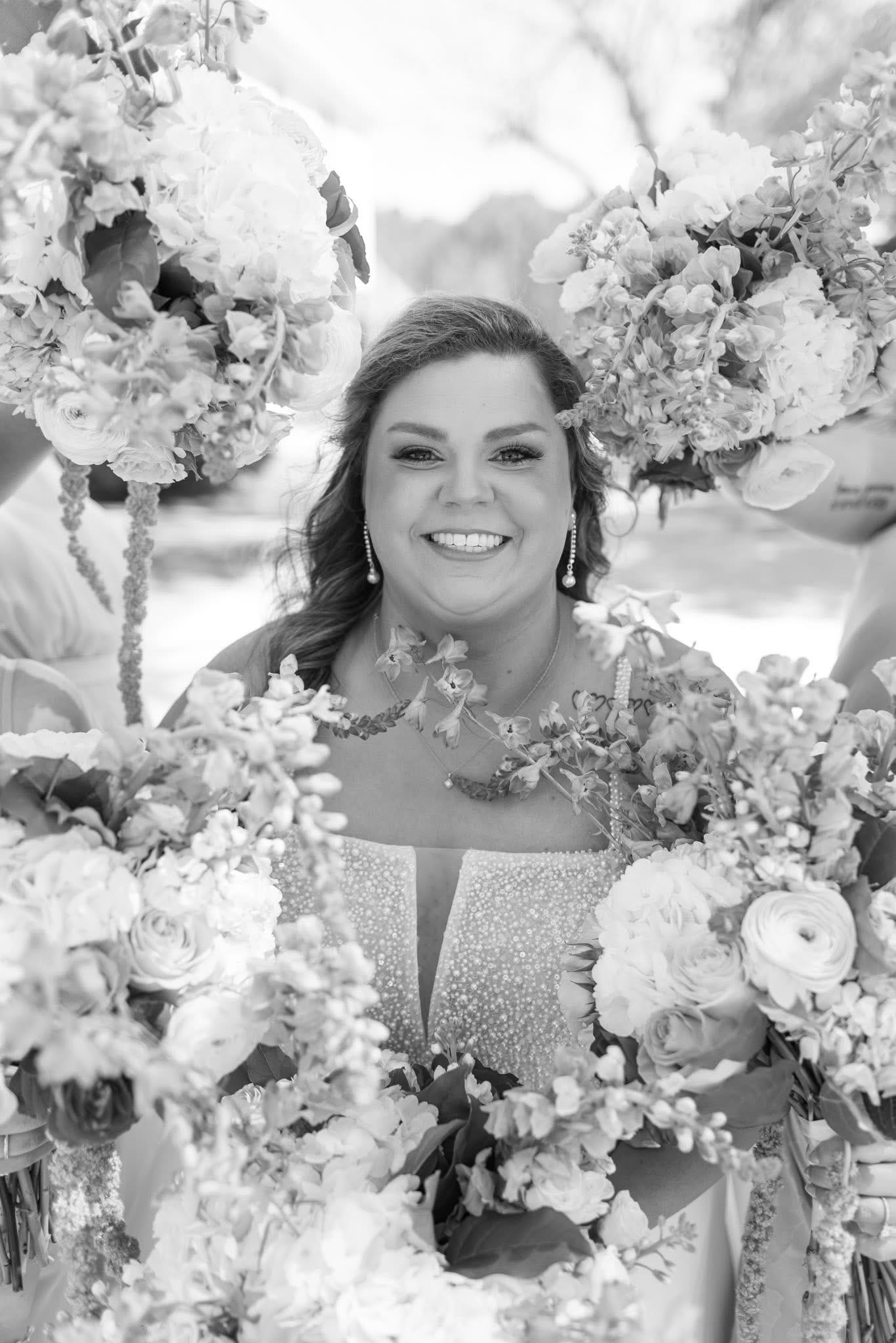 Black-and-white portrait of a smiling bride in a beaded gown surrounded by bridesmaids holding lush floral bouquets at Scarborough House wedding venue.