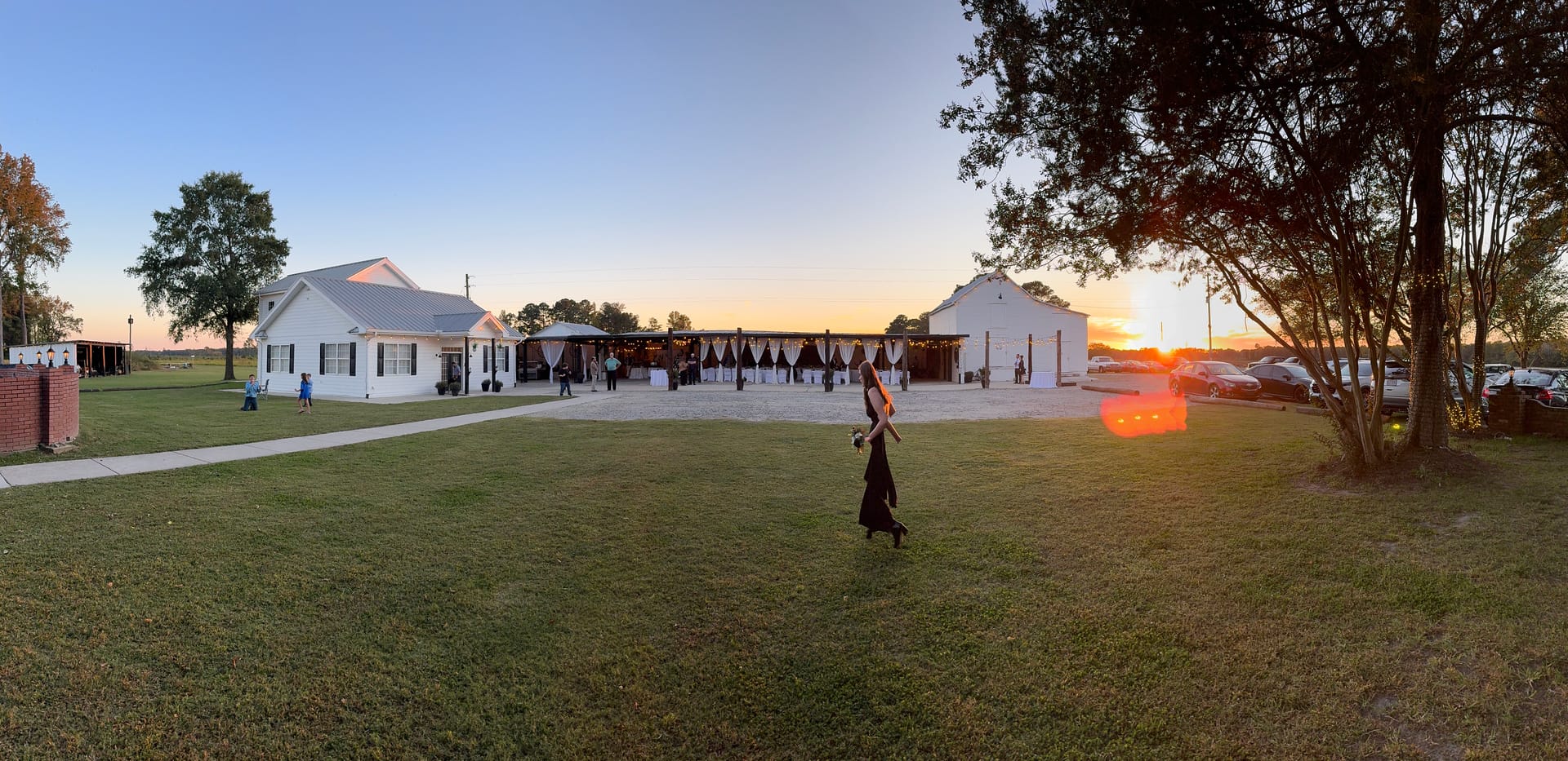 Panoramic golden-hour view of Scarborough House historic estate wedding venue at sunset, with guests gathered between the white manor house and grand hall as warm orange light glows across the ceremony lawn.