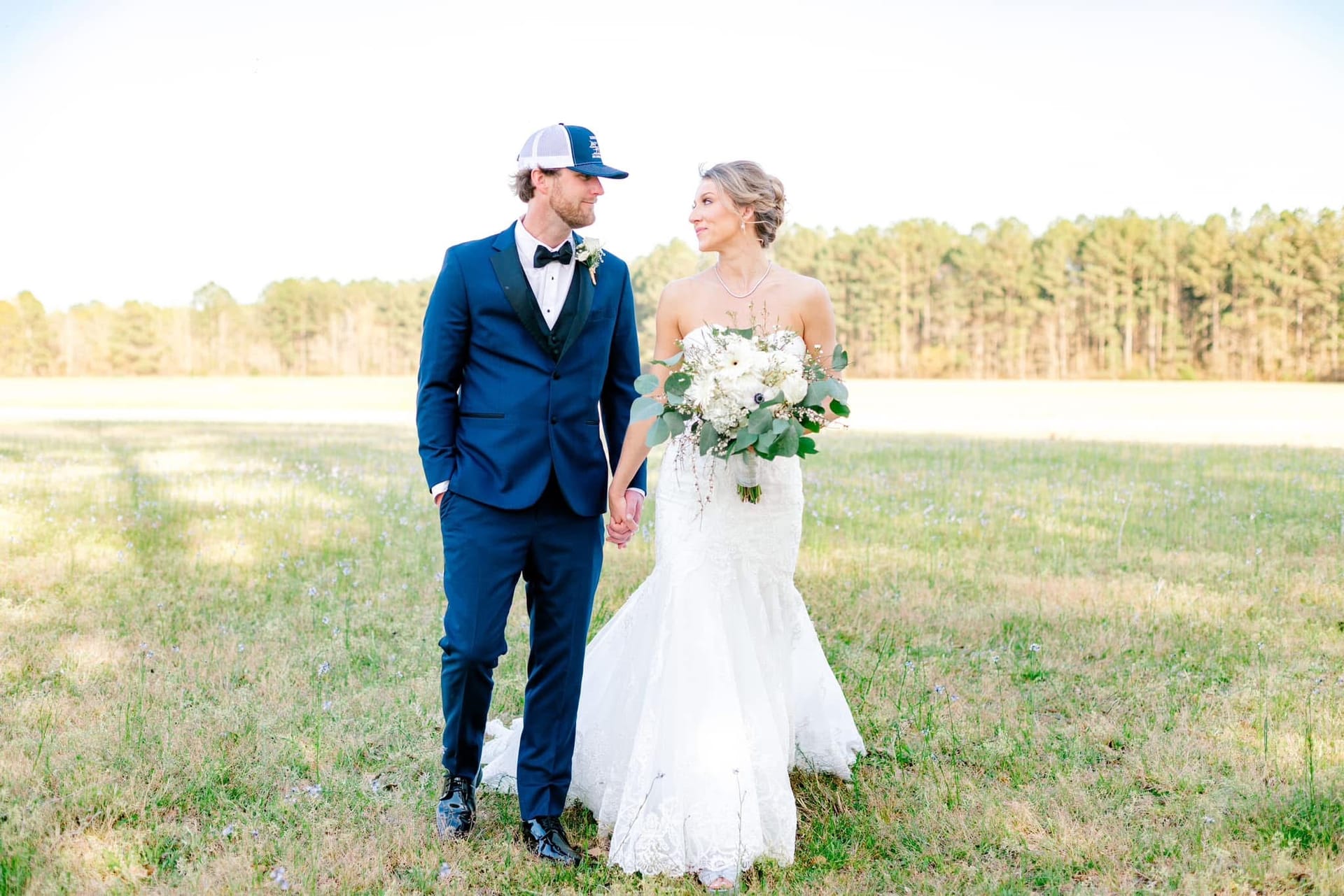 Bride in a strapless lace mermaid gown holding a white and greenery bouquet walks hand in hand with groom in a navy suit and trucker cap across an open field at Scarborough House estate during golden hour.