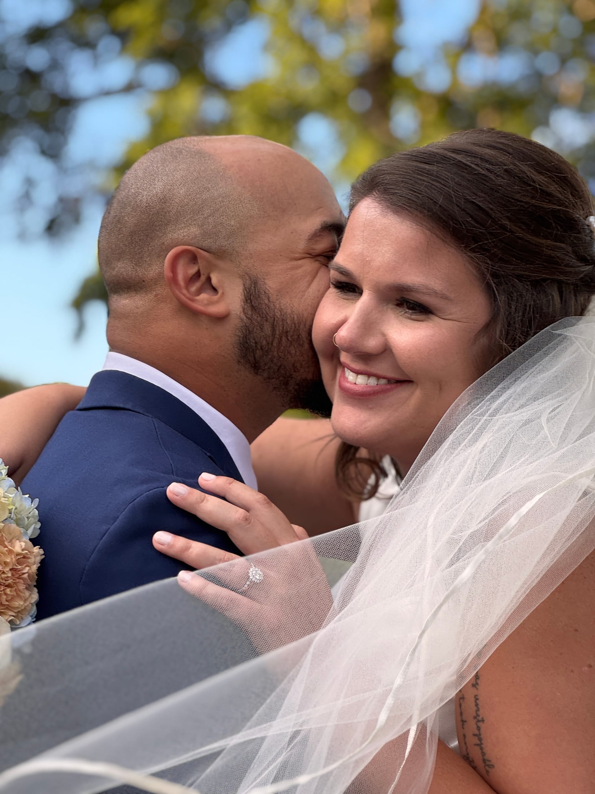 Groom in a navy suit kisses the smiling bride on the cheek as she holds her bouquet beneath oak trees at Scarborough House.