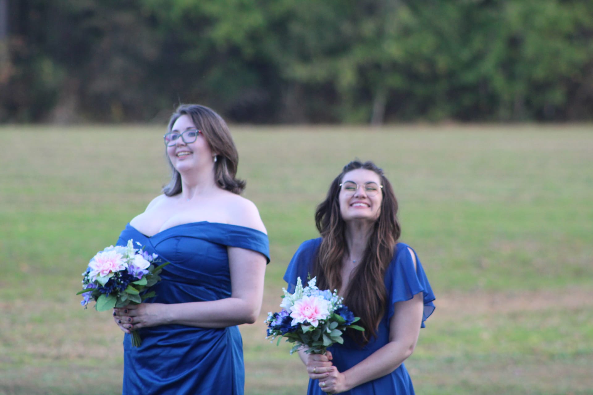 Two smiling bridesmaids in royal blue off-shoulder and flutter-sleeve gowns holding pink and blue bouquets stand on the ceremony lawn at Scarborough House with a lush green field and tree line behind them.