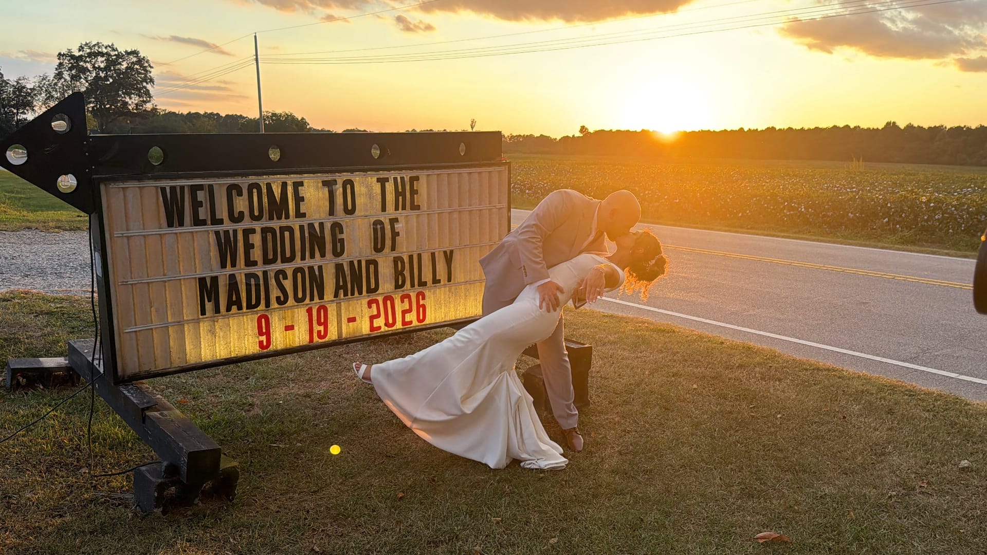 Bride and groom share a romantic dip kiss in front of an illuminated marquee wedding welcome sign at golden hour with a glowing sunset over open farmland fields behind them.
