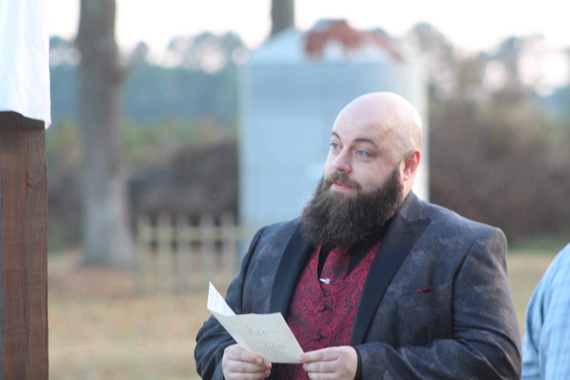 A bearded officiant in a dark blazer and red paisley shirt reads from a paper during an outdoor wedding ceremony at Scarborough House historic estate.
