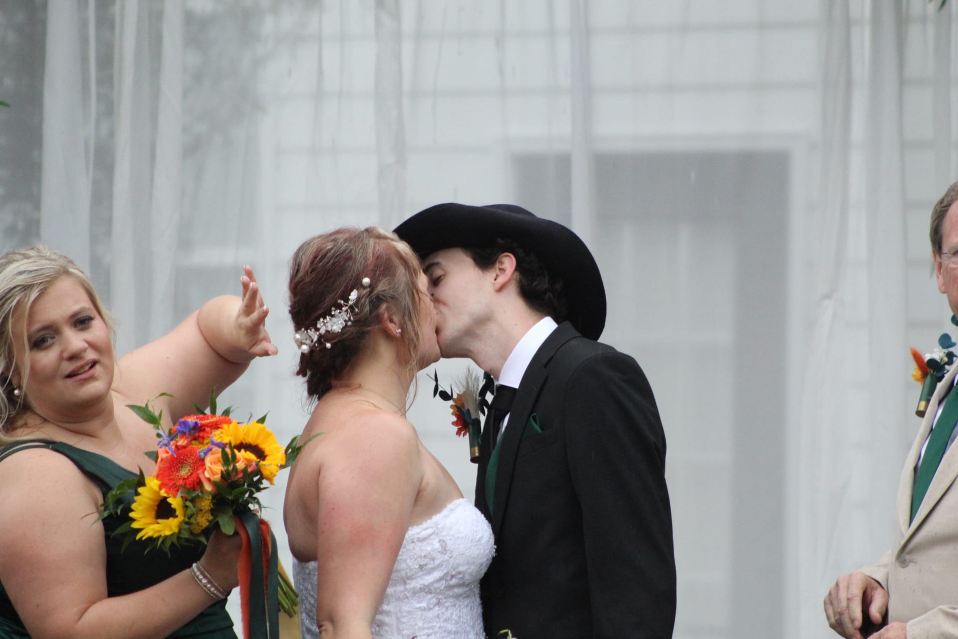 Bride and groom share their first kiss during an outdoor wedding ceremony at Scarborough House as a bridesmaid in emerald green holds a vibrant sunflower bouquet beside them.