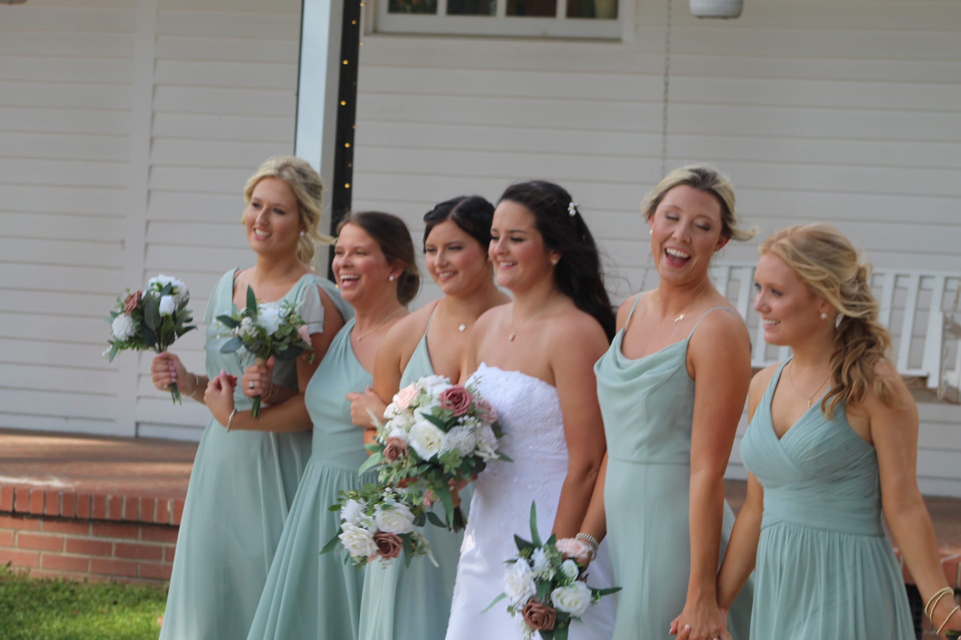 Bride in a white strapless gown laughing with five bridesmaids in sage green dresses holding floral bouquets in front of the historic white manor house at Scarborough House.