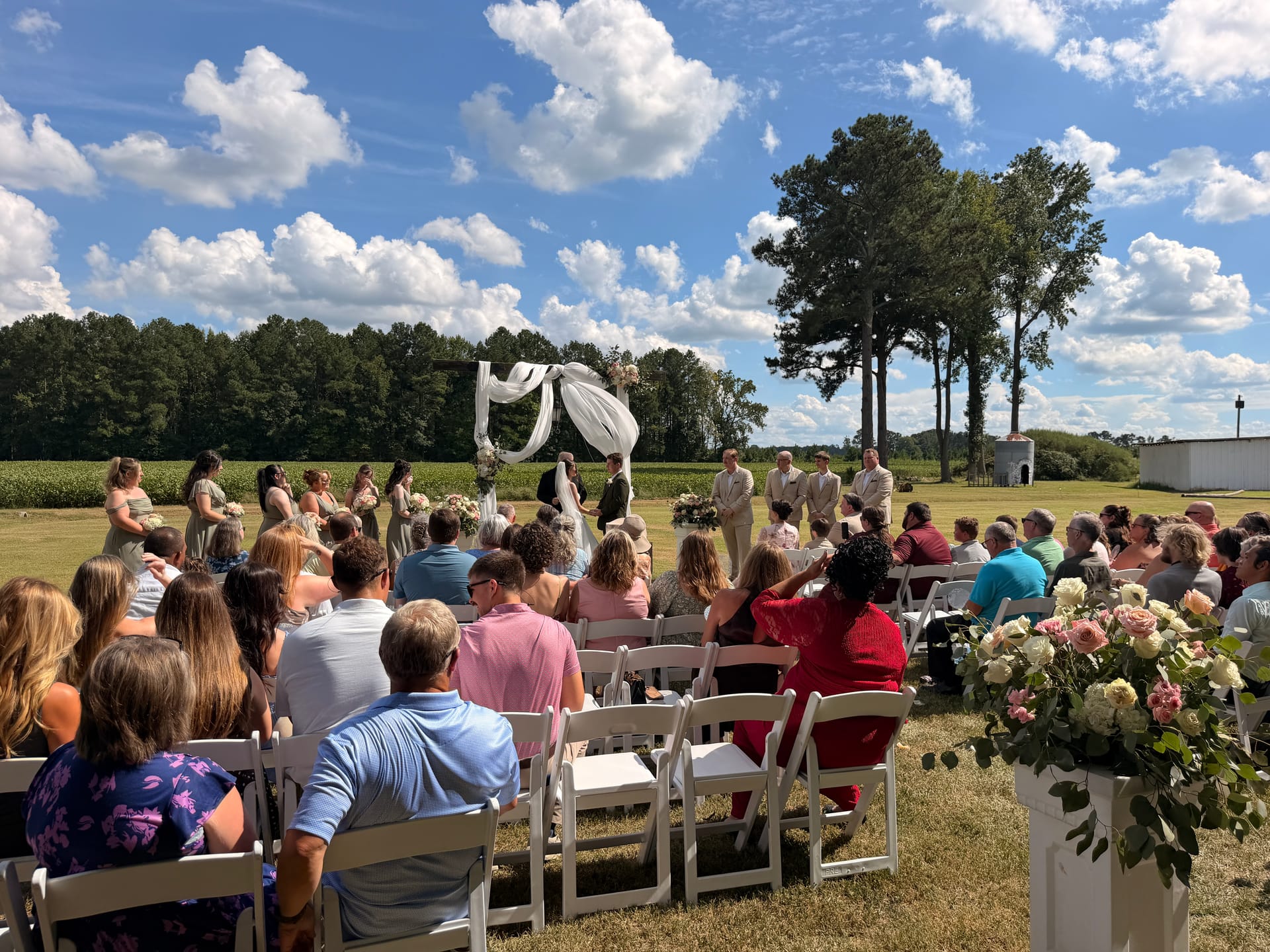 Guests seated in white chairs watch an outdoor wedding ceremony beneath a draped floral arch on the sun-drenched ceremony lawn at Scarborough House, surrounded by farmland fields and tall pines under a blue sky.