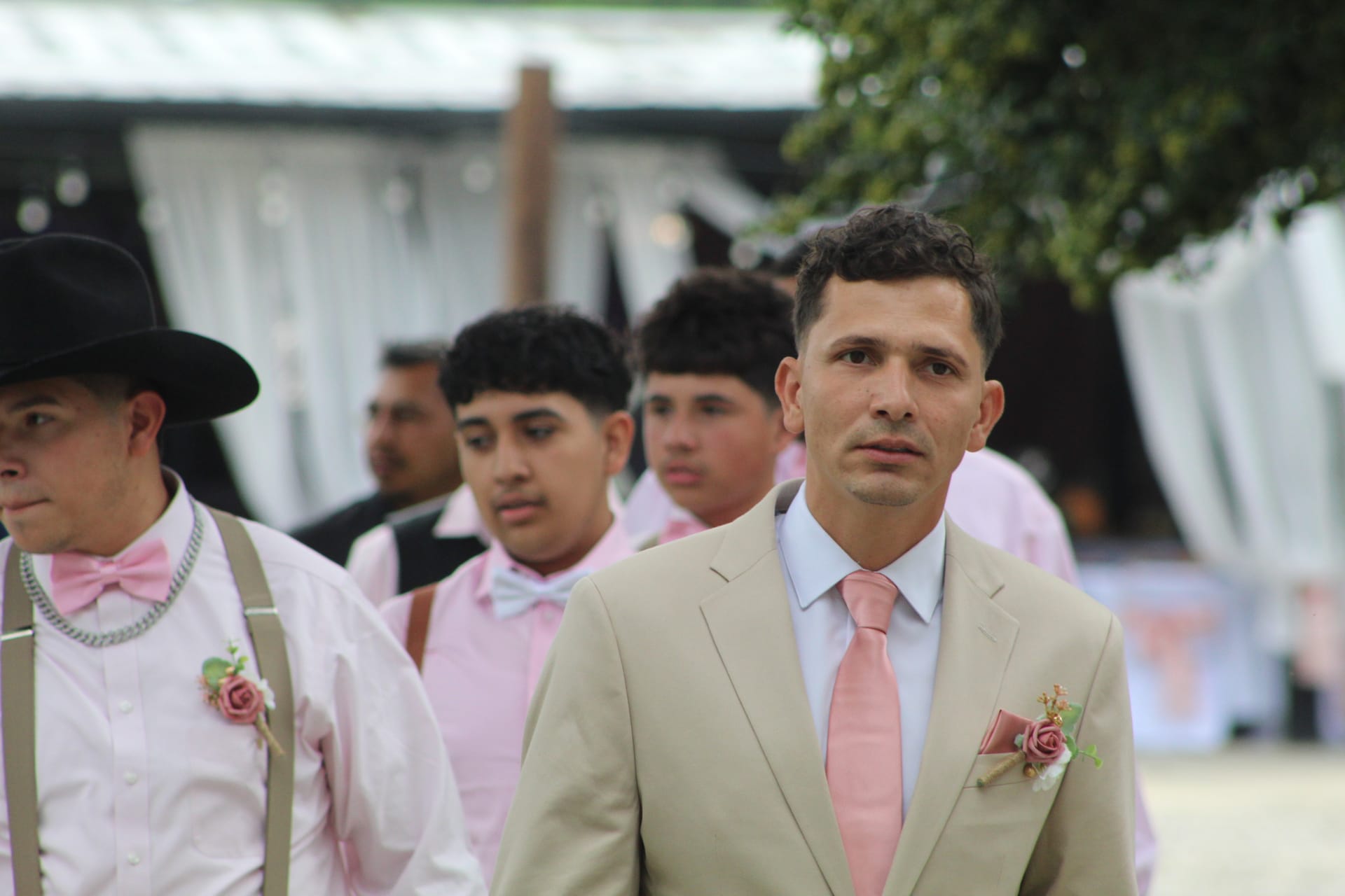 Groom in a tan suit with a pink tie and boutonniere leads his groomsmen dressed in pink bow ties and suspenders during an outdoor wedding processional at a historic estate venue.