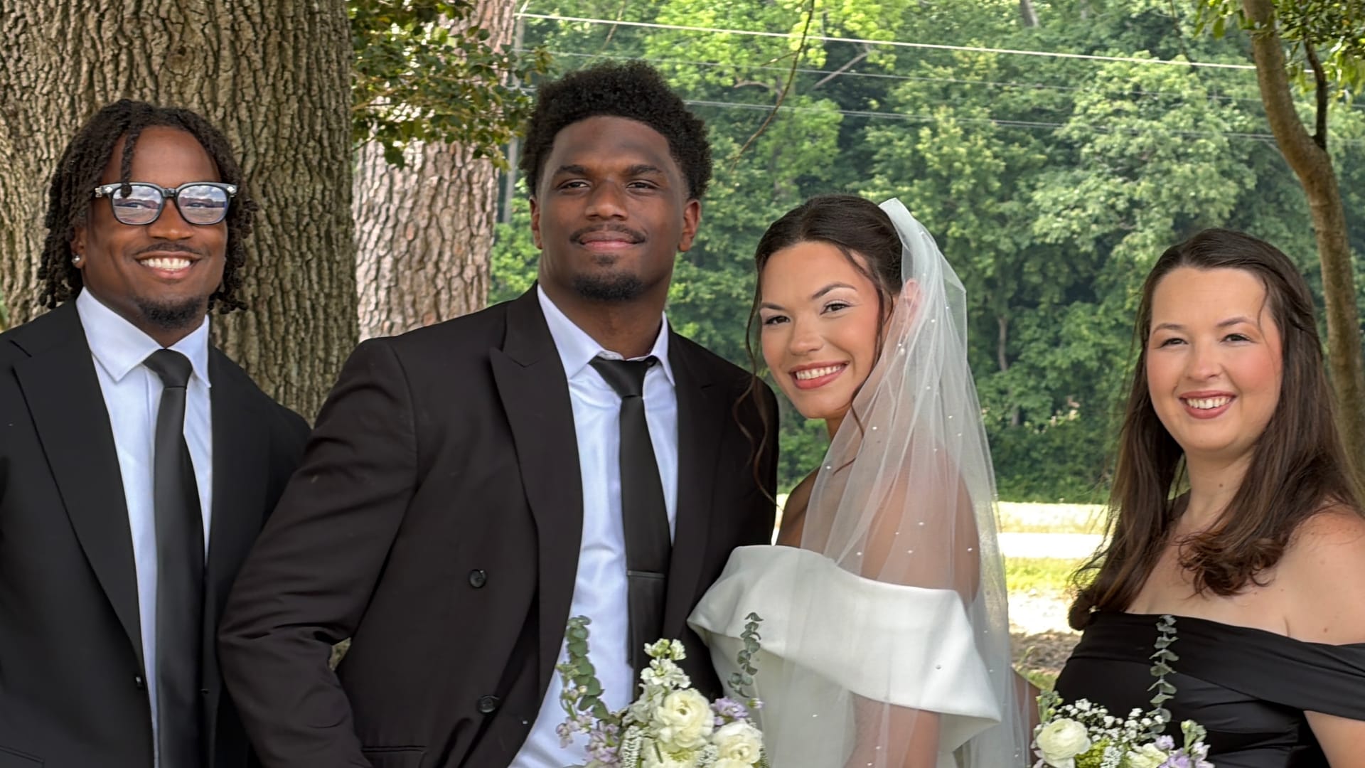 Bride in white off-shoulder gown and veil holding a white floral bouquet poses and smiles with three wedding guests in formal attire beneath large oak trees at Scarborough House.