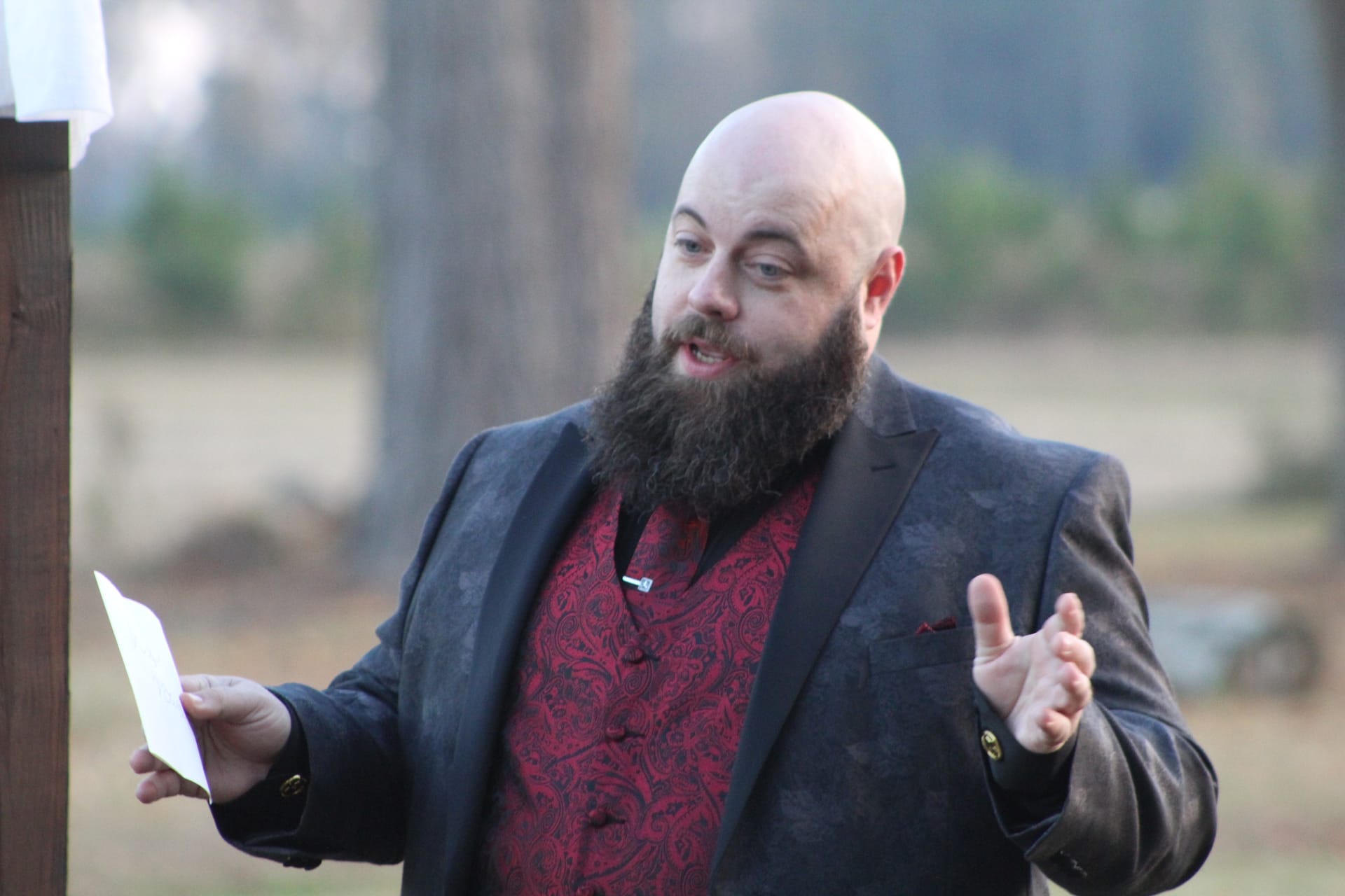A bald bearded officiant in a black blazer and red paisley shirt gestures expressively while speaking during an outdoor wedding ceremony at Scarborough House.