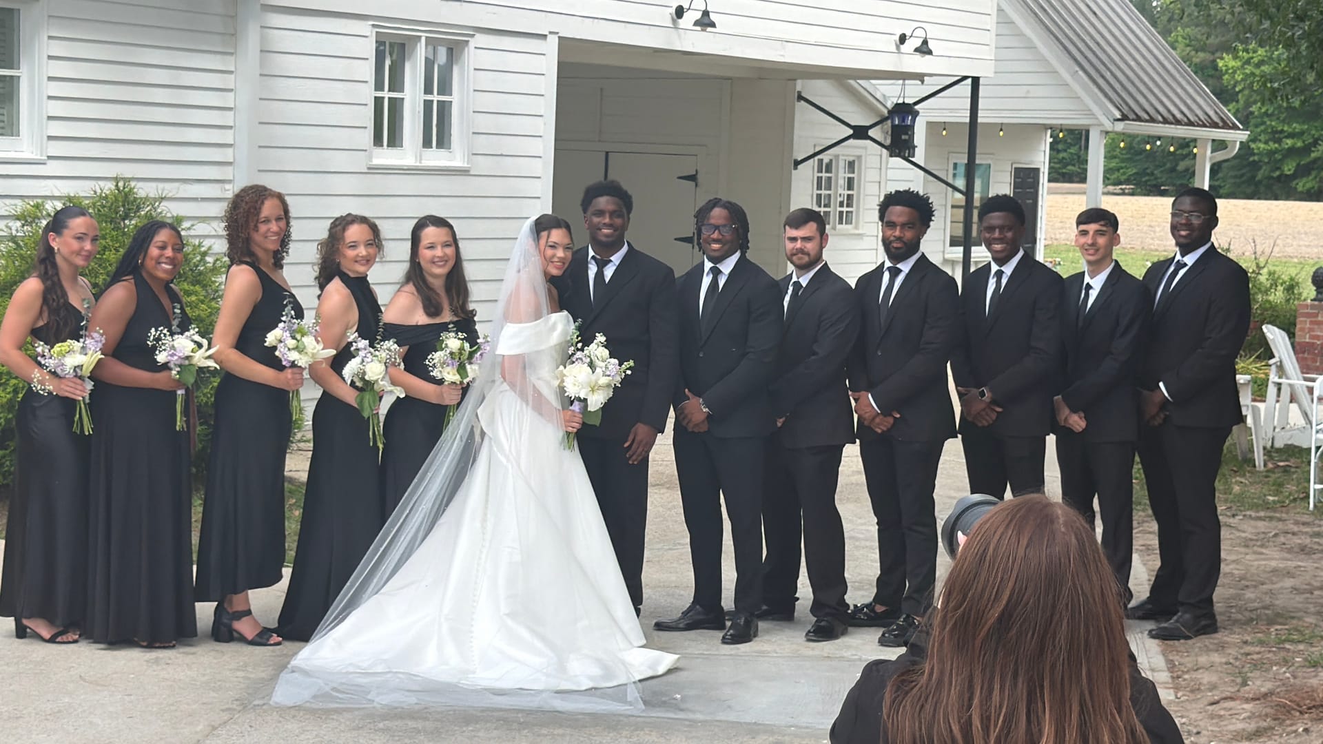Bride in white gown and veil poses with bridesmaids in black dresses and groomsmen in black suits in front of a historic white clapboard building at Scarborough House.