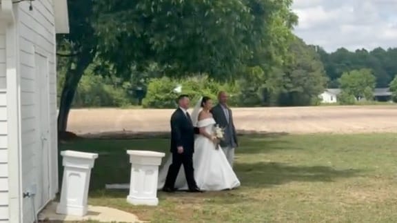 Bride in a white off-shoulder gown walks with two escorts across the sunlit ceremony lawn at Scarborough House, with lush oak trees and open farmland fields in the background.