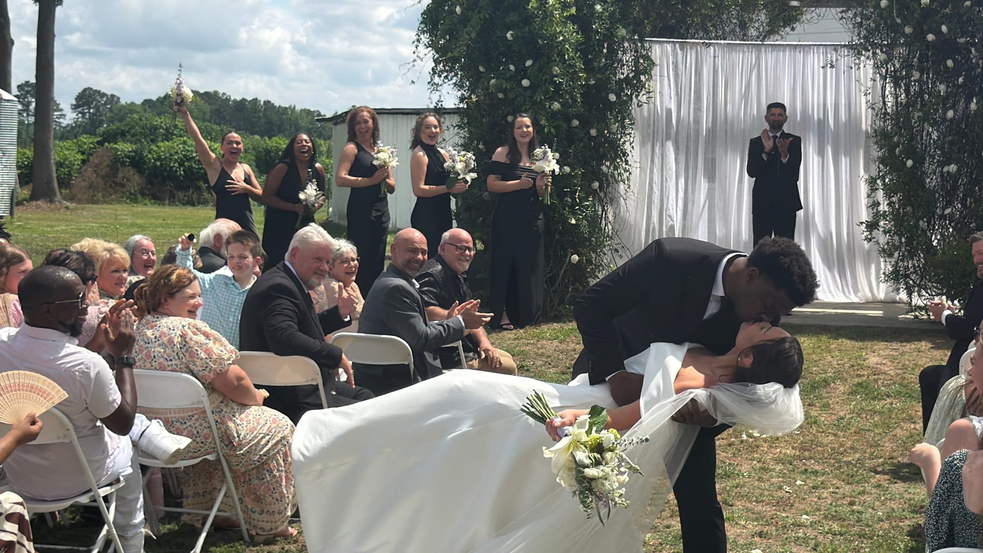 Groom dips his bride for a dramatic kiss while wedding guests and bridesmaids cheer during an outdoor ceremony at a historic estate venue.