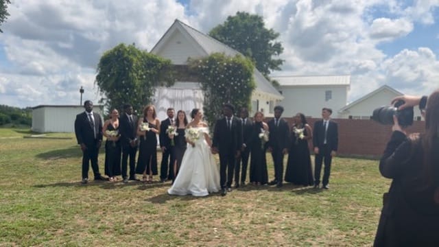 Bride and groom stand with their full wedding party dressed in black in front of a vine-covered white chapel arch on the ceremony lawn at Scarborough House.