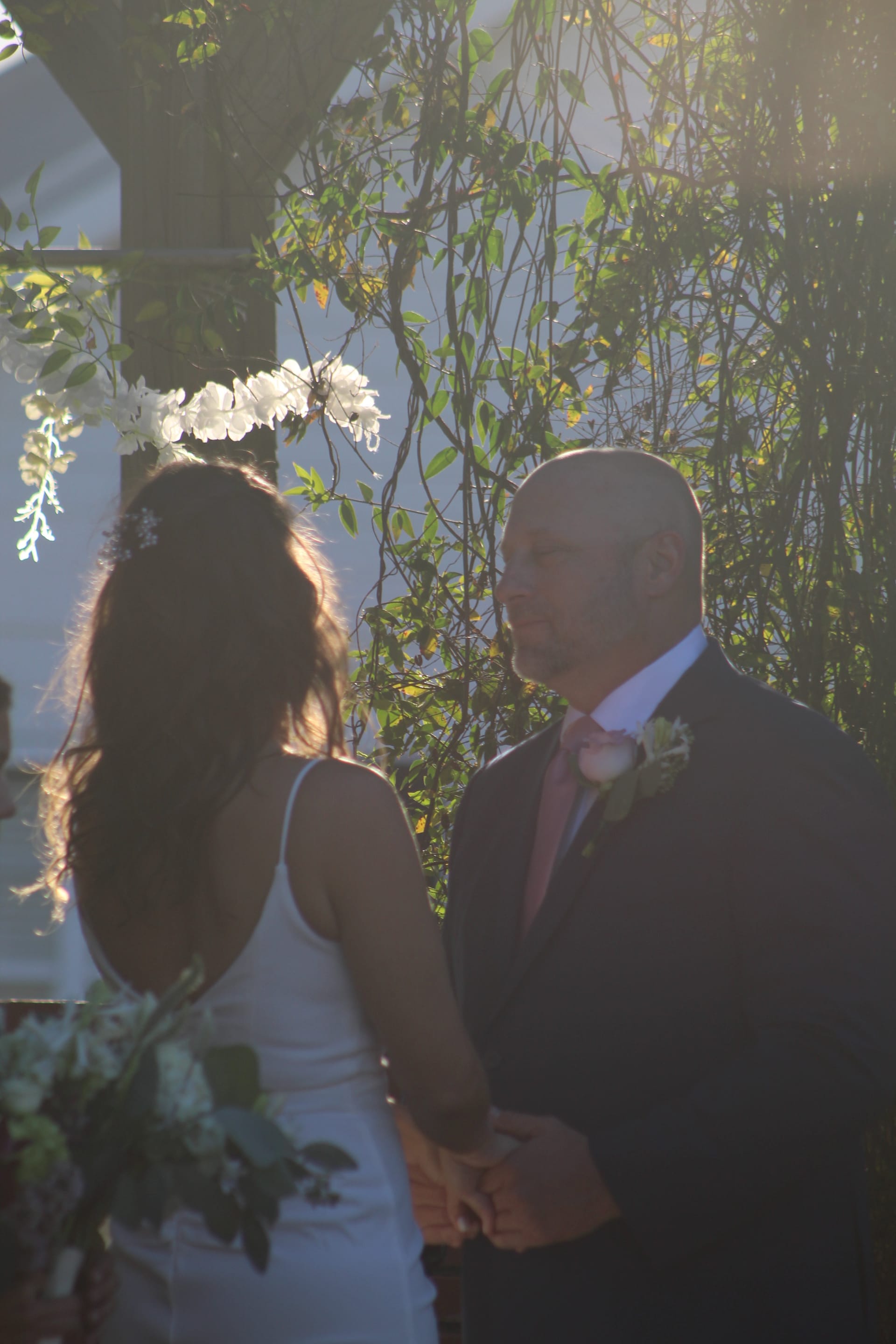 Bride and groom hold hands facing each other beneath a flowering vine arbor during an outdoor garden wedding ceremony bathed in warm golden-hour light.