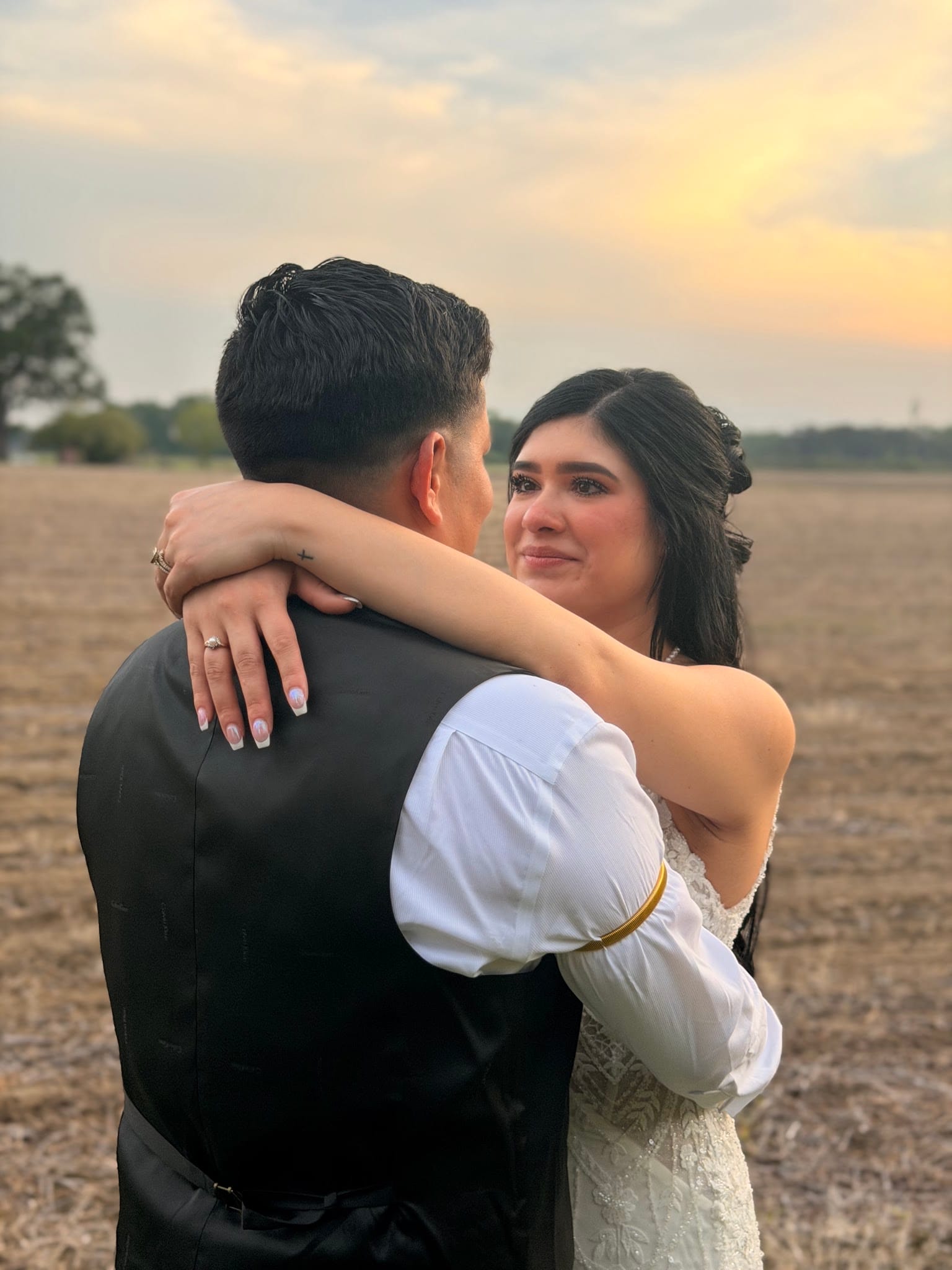 Tearful bride in a lace gown wraps her arms around her groom at sunset over open farmland fields at Scarborough House wedding venue.