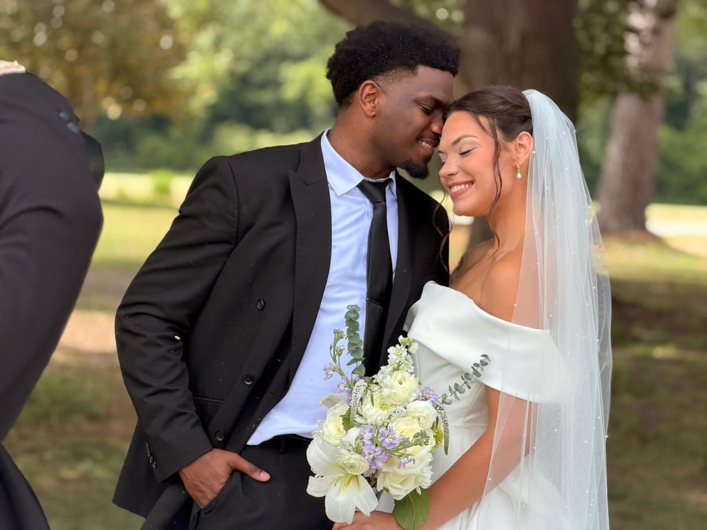 Bride in an off-shoulder white gown and veil smiles as groom in a black suit leans his forehead toward hers beneath towering oak trees at Scarborough House estate.