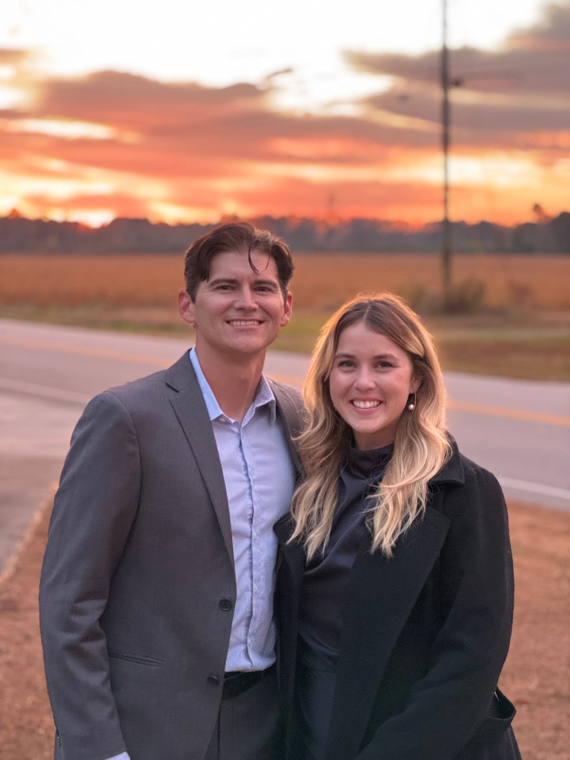 A smiling couple dressed in formal attire poses together on a rural road at Scarborough House with a vibrant orange and pink sunset glowing over the surrounding farmland fields.