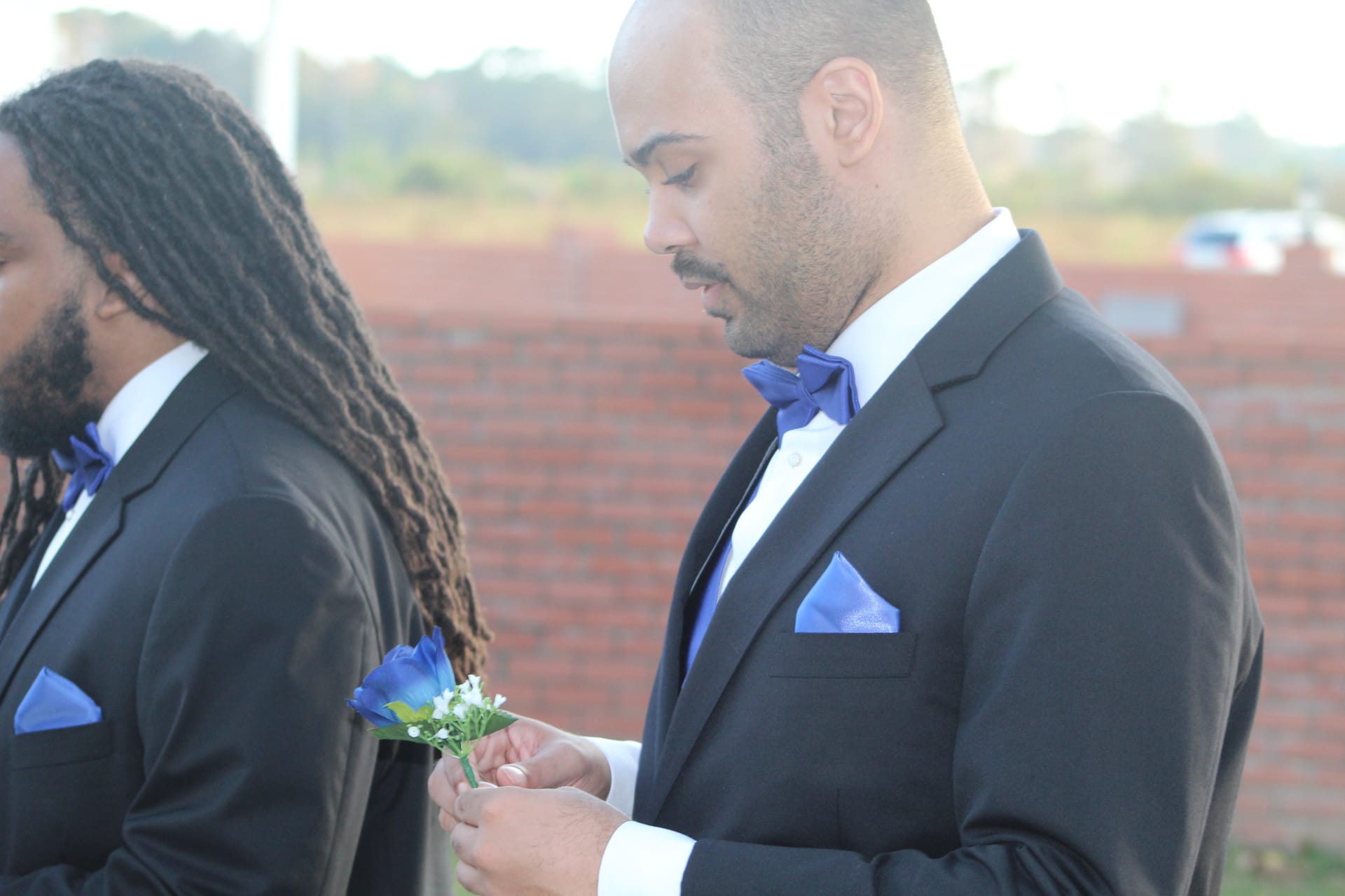 Two groomsmen in black tuxedos with royal blue bow ties and pocket squares prepare boutonniere outside a historic brick estate venue.