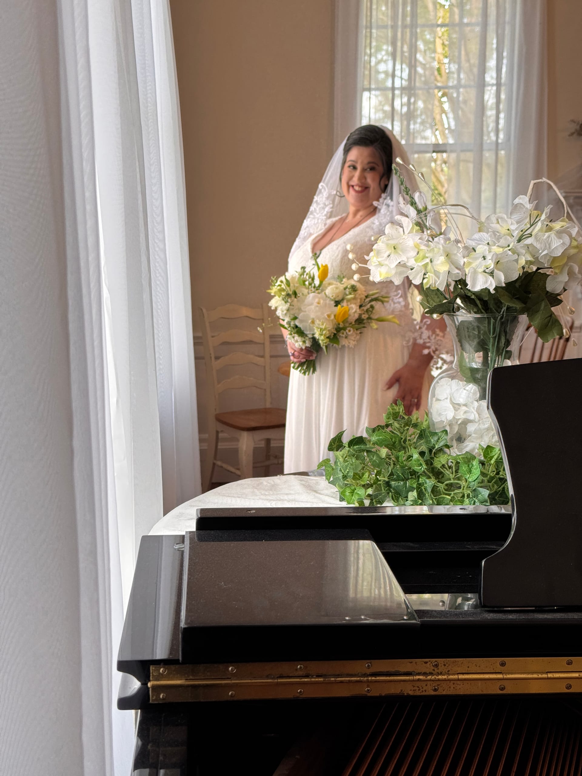 A smiling bride in a white lace gown and veil holds a bouquet of white and yellow flowers while standing near a window in the grand hall of Scarborough House, with a black grand piano and white floral arrangement in the foreground.