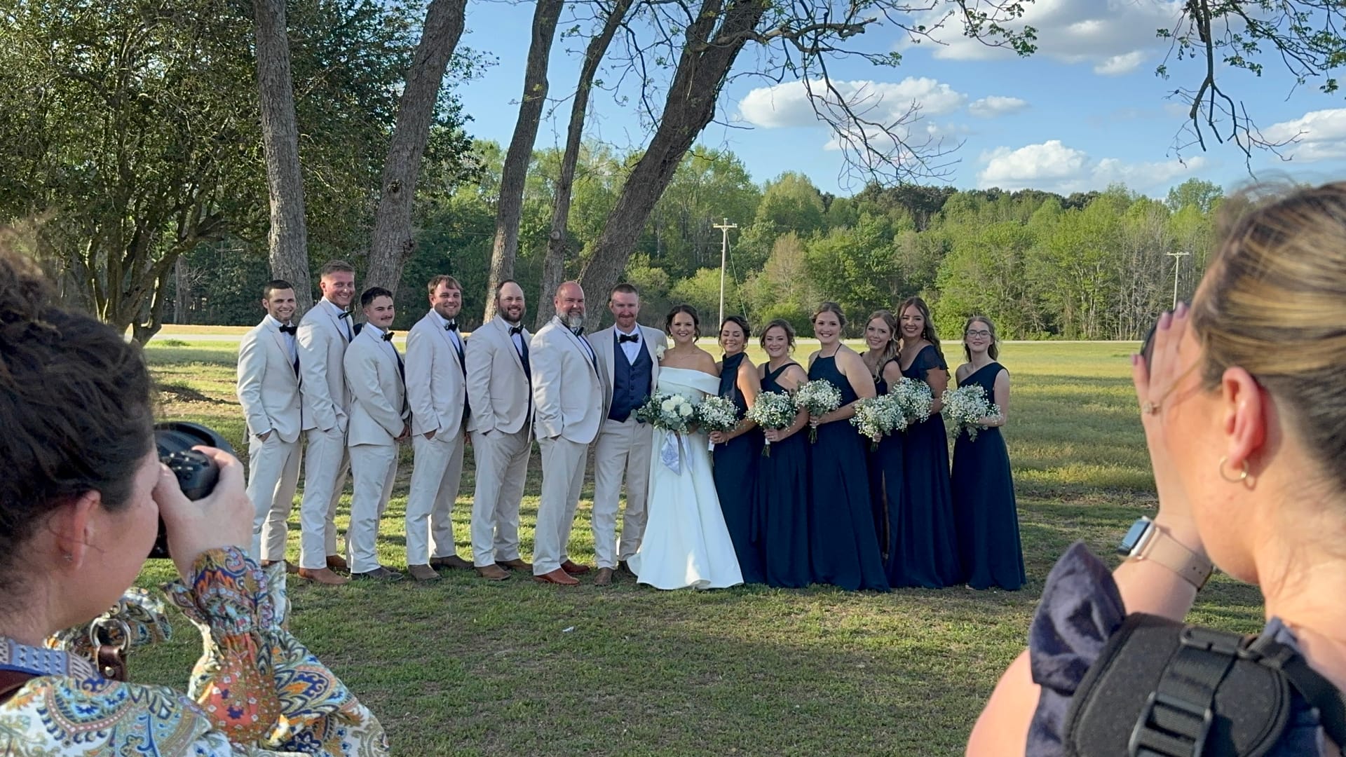 Full wedding party portrait under mature oak trees at Scarborough House with groomsmen in ivory suits and bridesmaids in navy gowns flanking the bride and groom on the ceremony lawn.