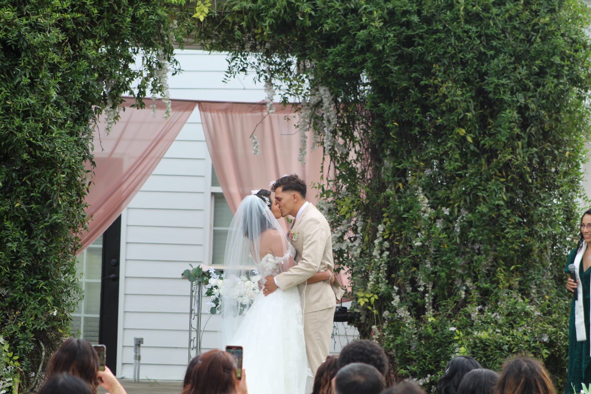 Bride and groom share their first kiss beneath a lush green archway adorned with blush pink drapery and white florals at an outdoor garden wedding ceremony.