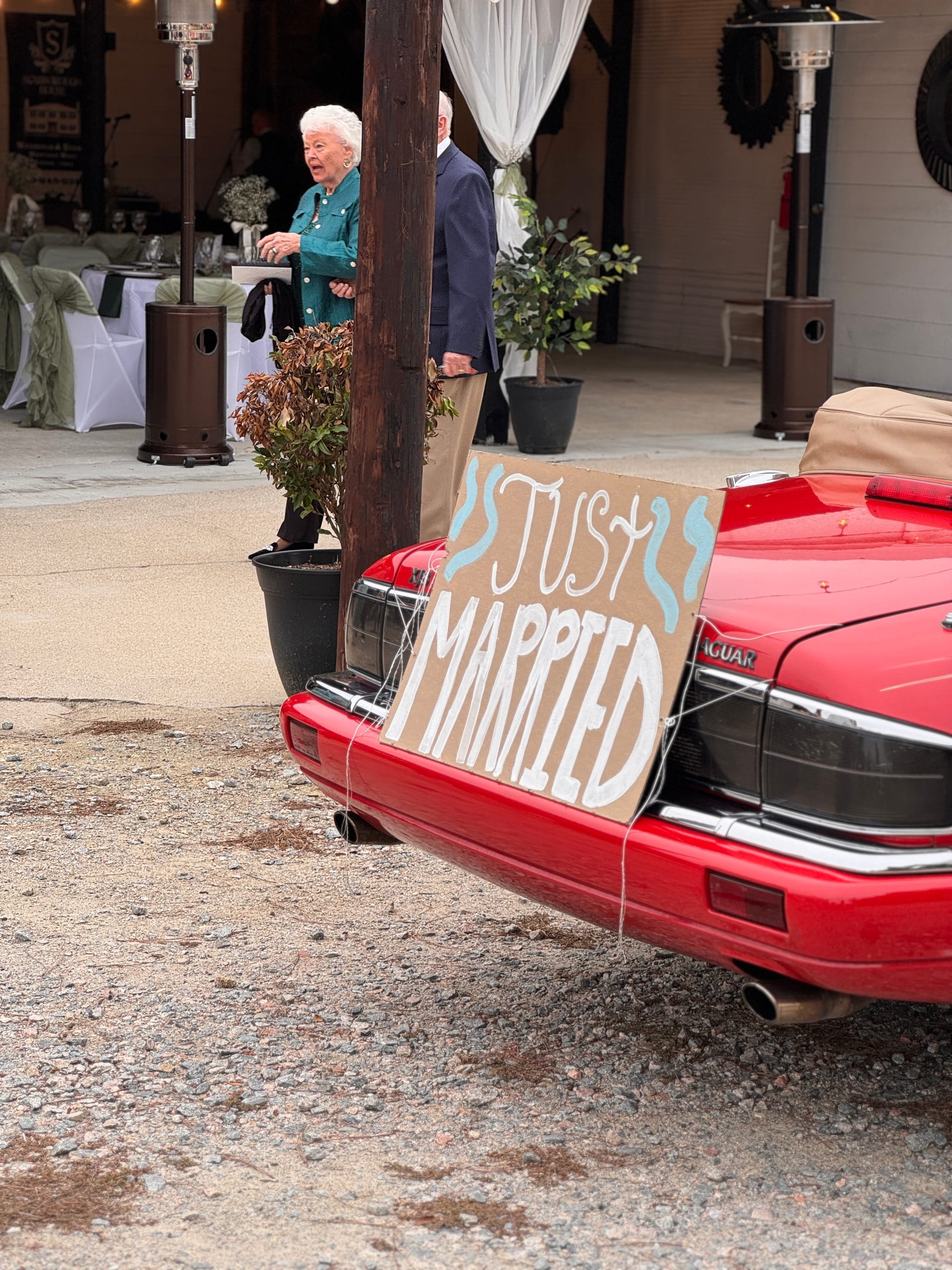 A handmade Just Married cardboard sign tied to the back of a classic red Jaguar convertible outside a wedding venue with guests visible in the background.