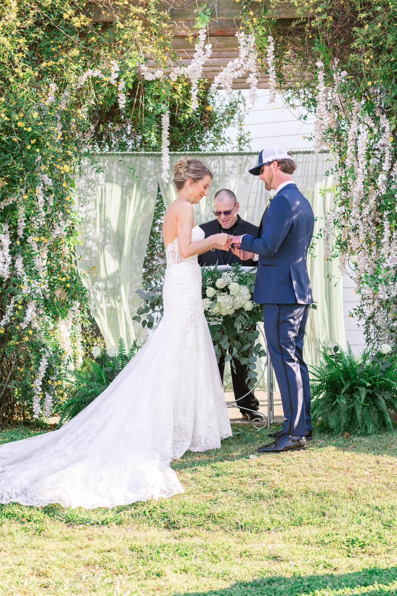 Bride placing a ring on the grooms finger during an outdoor wedding ceremony beneath a floral and greenery draped arch at Scarborough House estate.